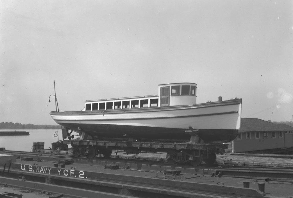 Black and white photo of a boat on a flat bed railcar marked with 'U.S. Navy YCF 2,' near a body of water, with a building and trees in the background.