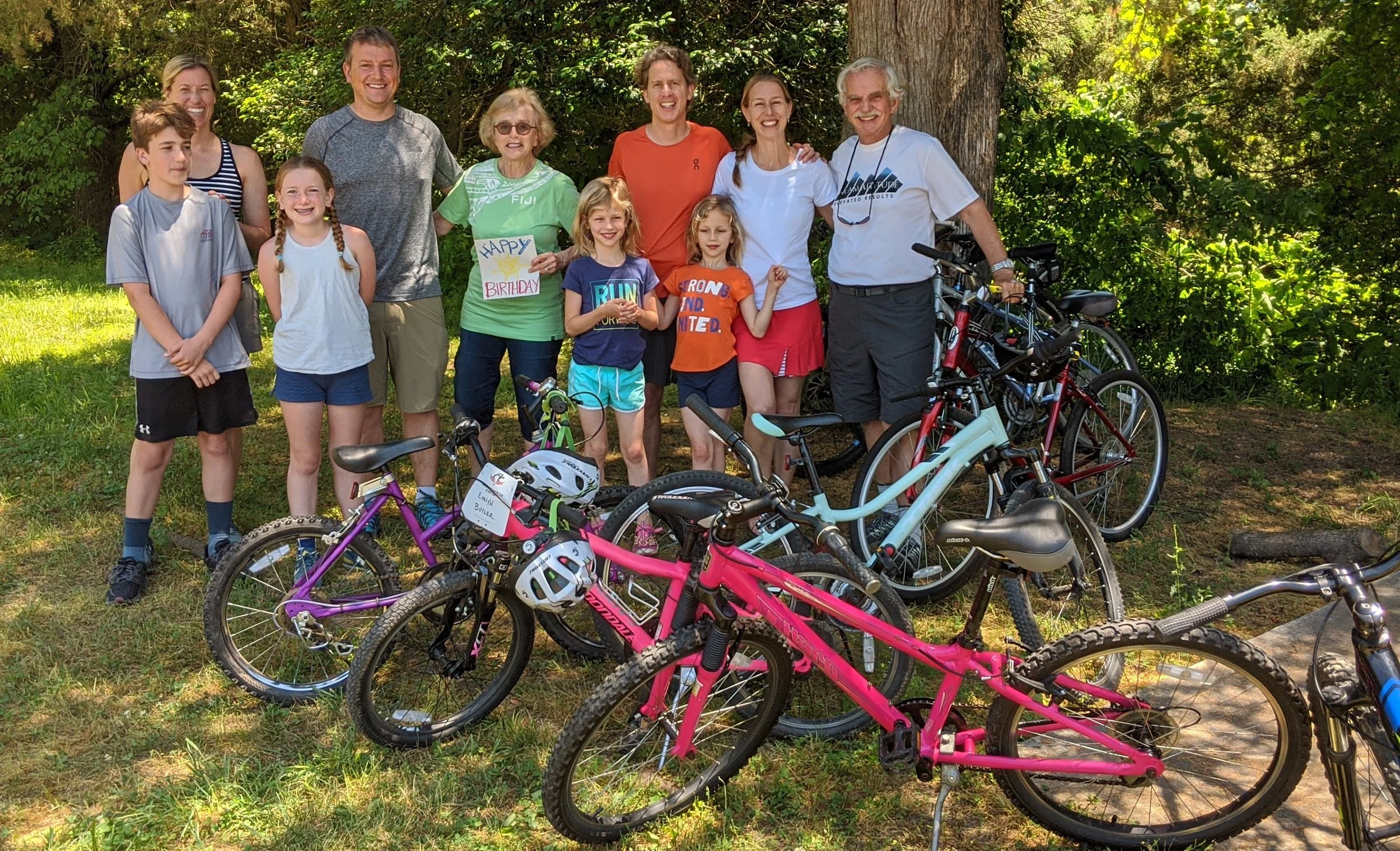 A group of people, including children and adults, gathered outdoors in front of trees, smiling. Several bicycles are parked in the foreground, and one person is holding a colorful sign that says "HAPPY BIRTHDAY."