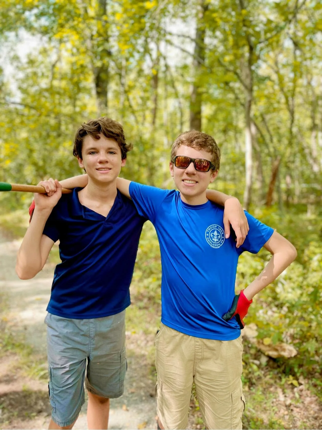 Two boys standing outdoors in a wooded area with trees, smiling, with their arms around each other. One boy holds a stick over his shoulder, and the other wears sunglasses. Both are wearing casual t-shirts and shorts.