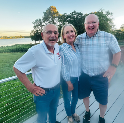 Three smiling people, two men and one woman, standing outdoors on a wooden deck near a body of water with trees in the background during the daytime.