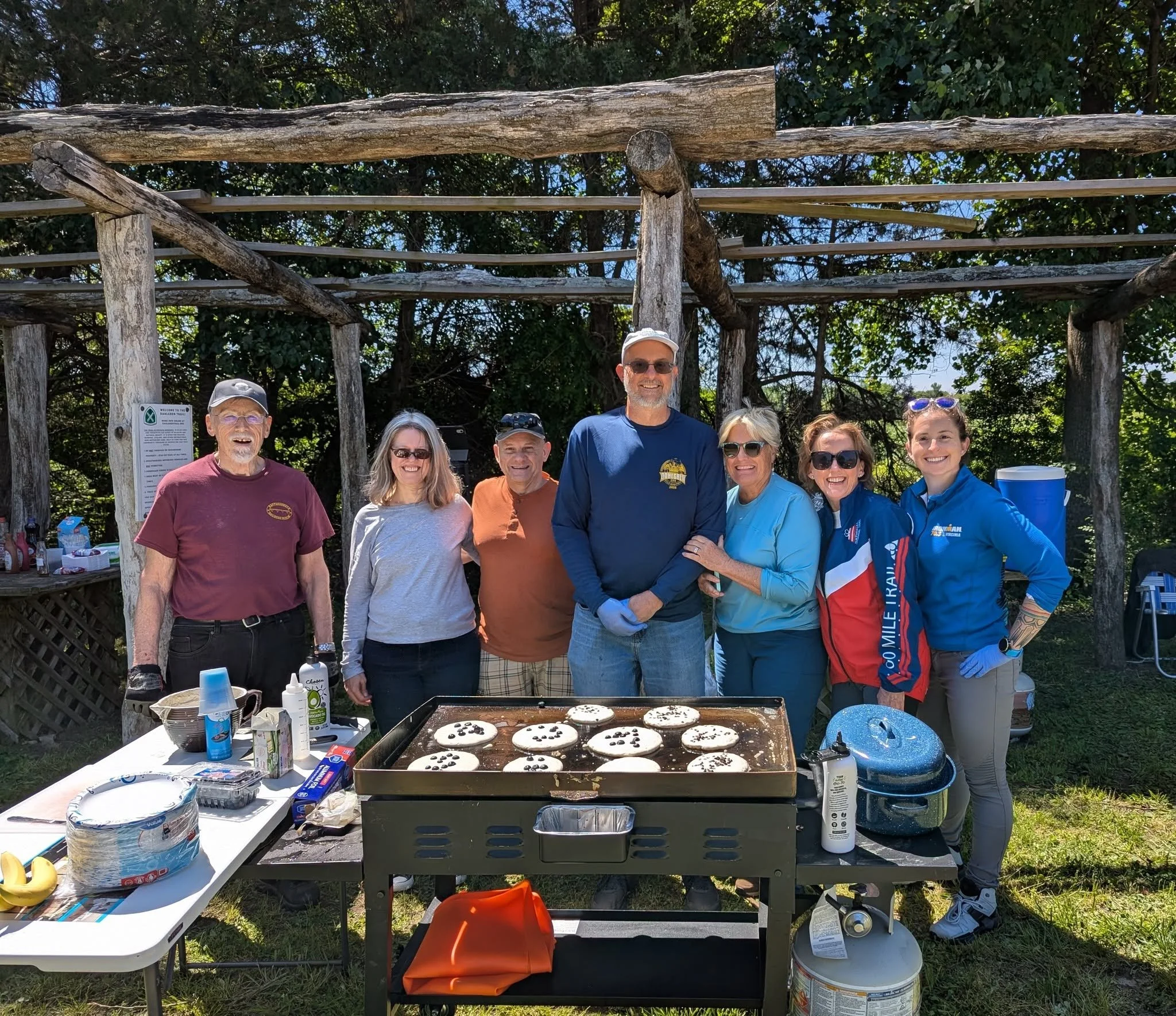 Group of people gathered outdoors under a wooden canopy with trees in the background, standing behind a grill with cookies, and table with food items and supplies during a sunny day.