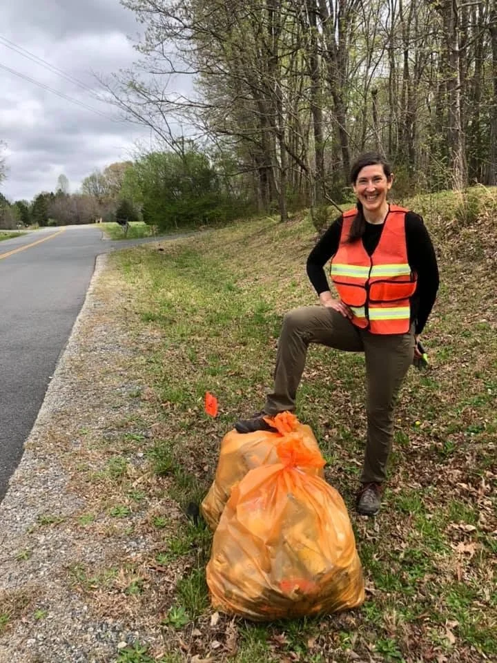 Woman wearing an orange safety vest and brown pants standing next to bags of collected trash on the side of a rural road.