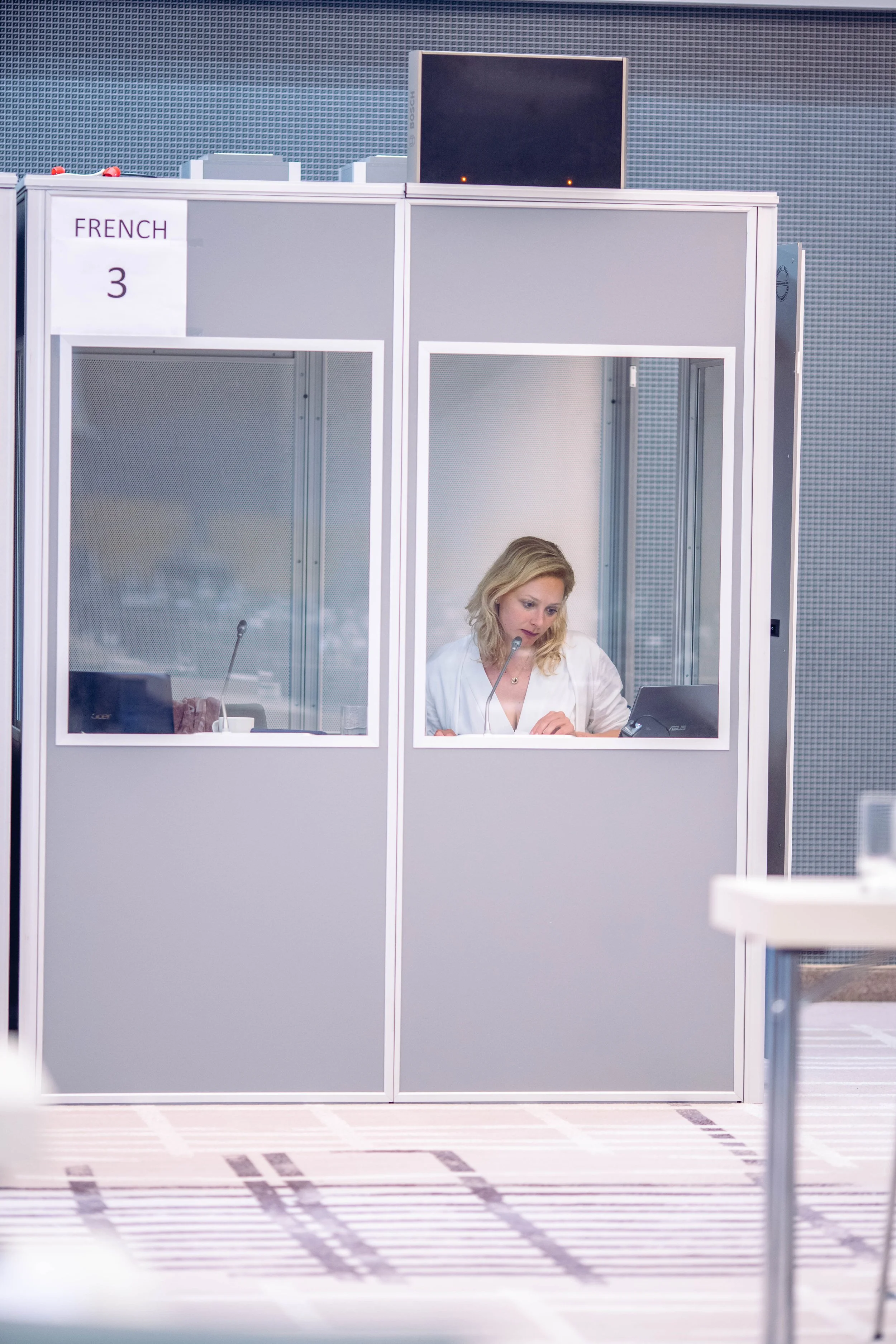 Eine Frau sitzt in einem kleinen, schallisolierten Raum mit Glasfenstern, arbeitet an einem Laptop und hat ein Mikrofon vor sich. Es ist eine Art Hör- oder Dolmetscherkabine im Flughafen.