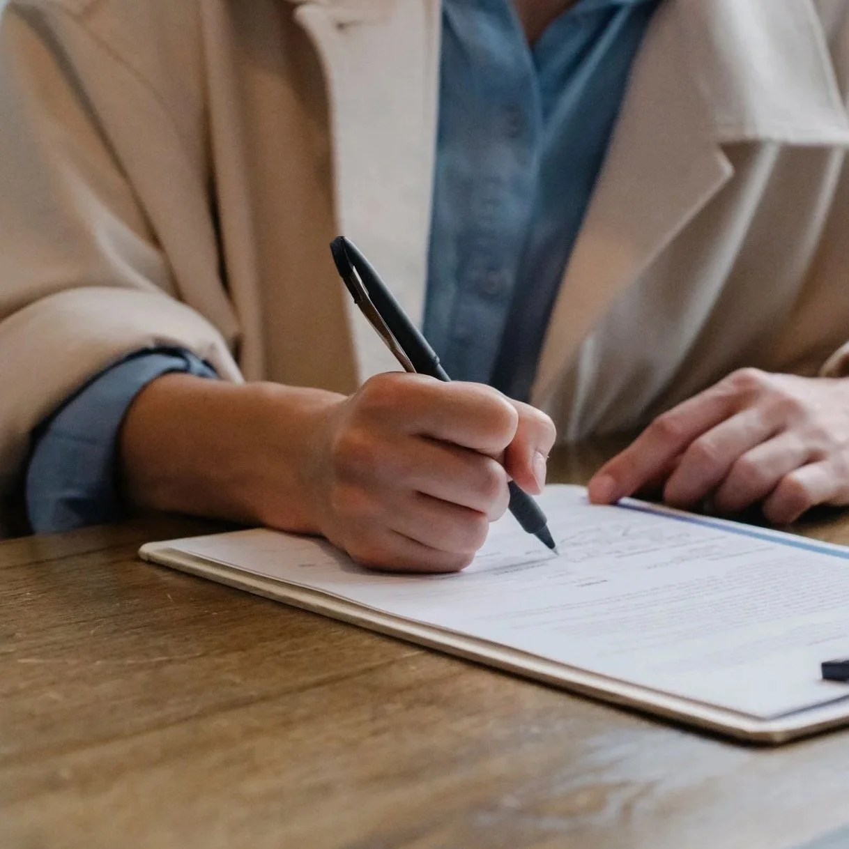 A light skin toned person wearing a beige coat and a blue shirt is writing in a notebook with a black pen on a wooden table.
