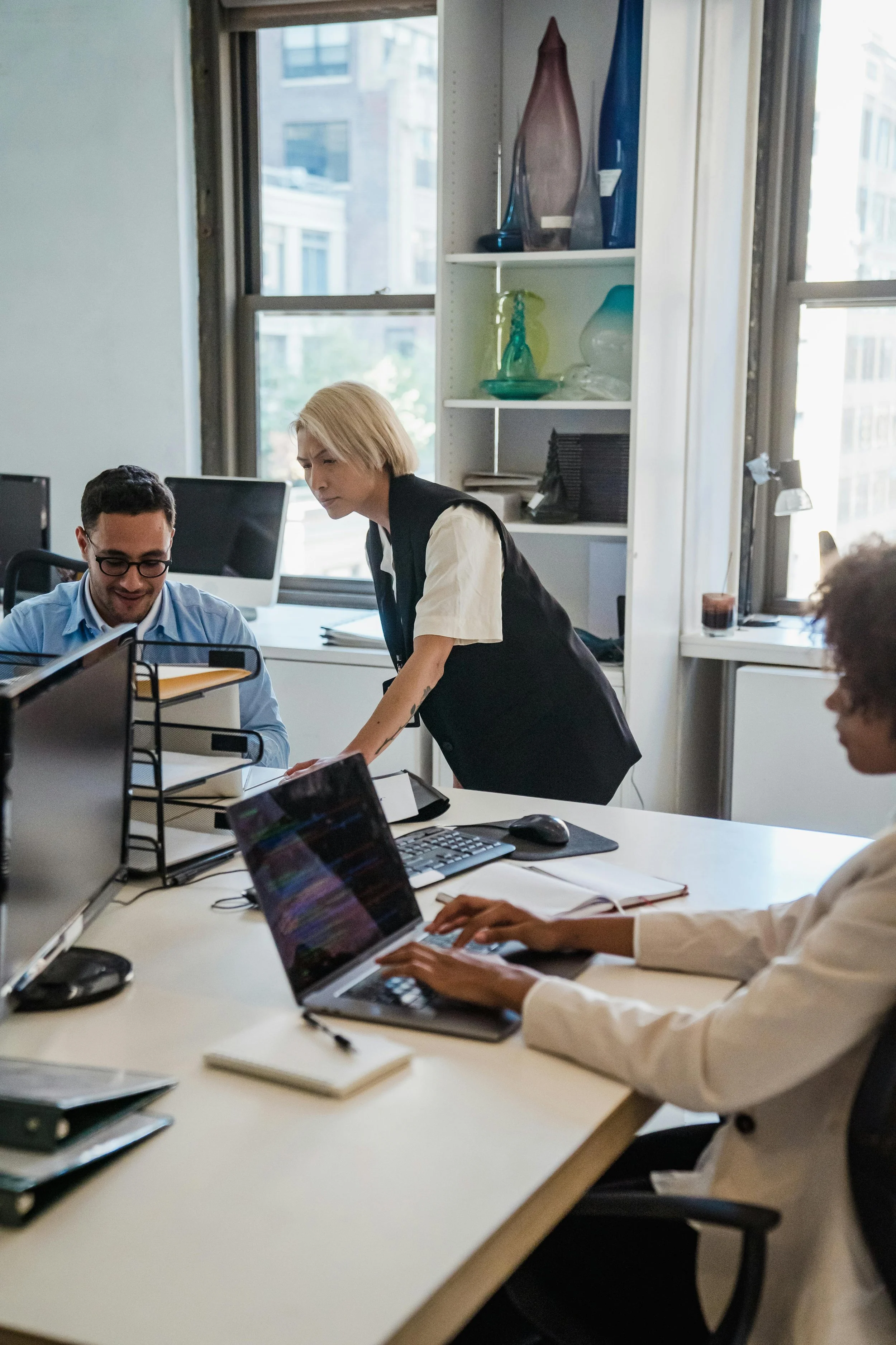 Office meeting with four diverse professionals working at a white table, using laptops and documents, in a modern office with large windows, shelves with decorative glass vases, and cityscape view outside.