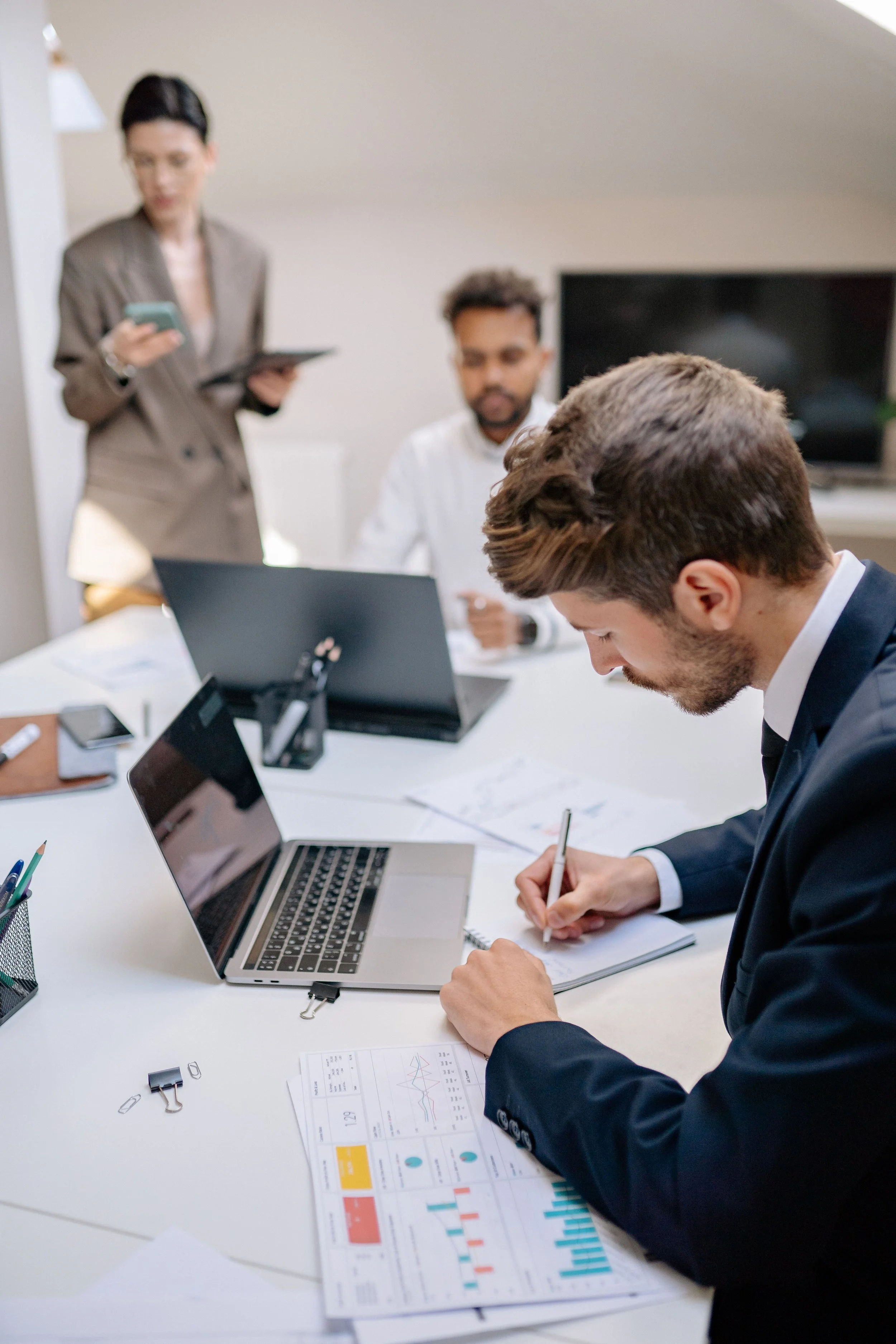 Business meeting with three people at a table, one person taking notes, another working on a laptop, and a woman standing in the background looking at her phone.