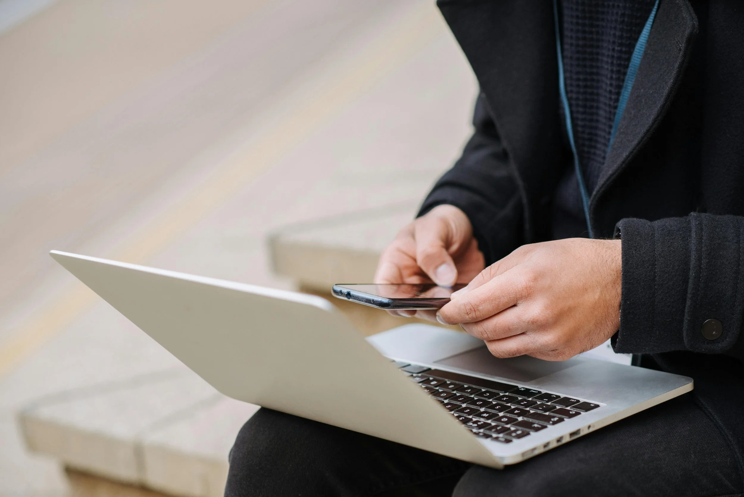 Person with a black jacket and blue trim using a smartphone while sitting in front of a laptop.