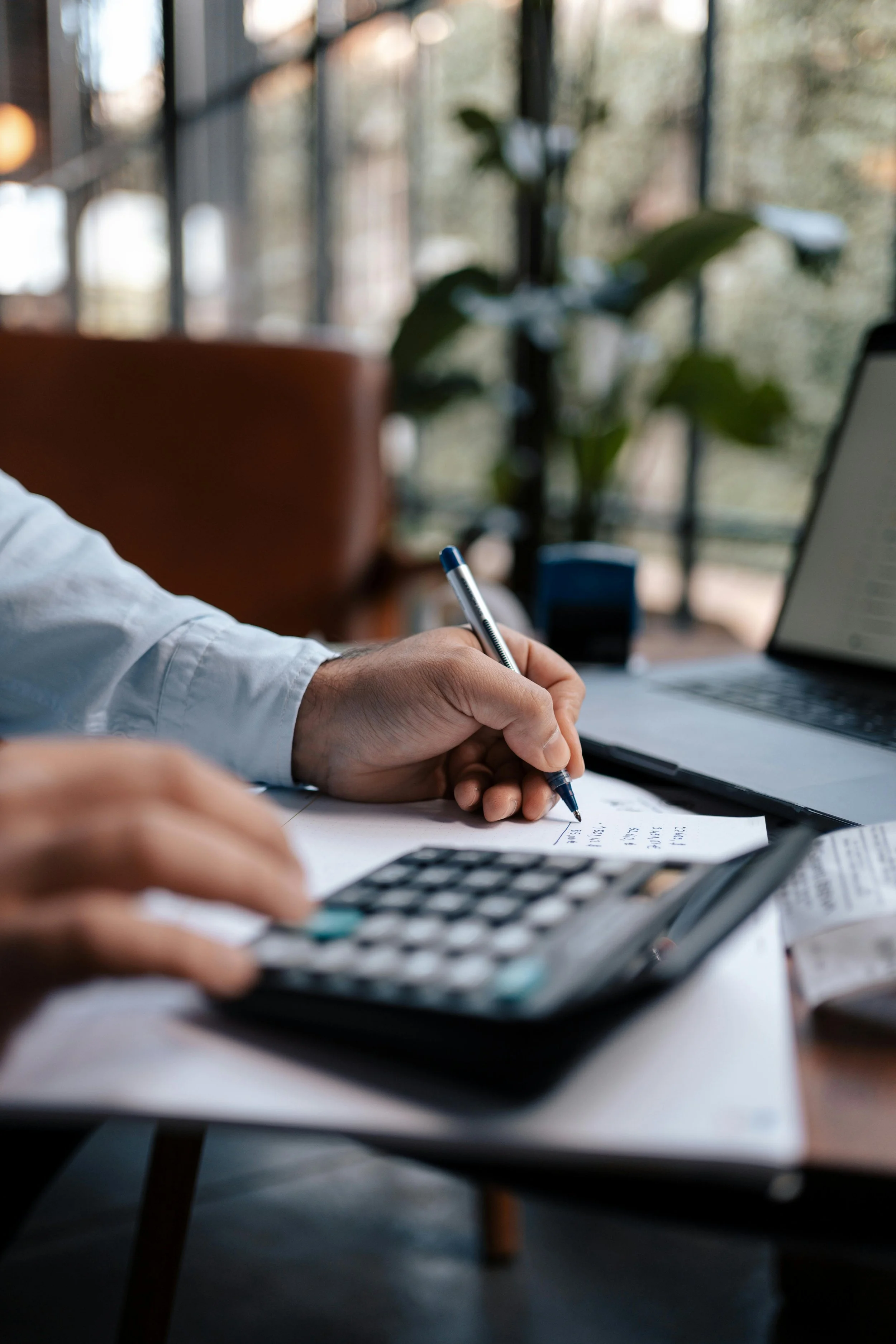 Close-up of a person's hand writing on a piece of paper at a desk, with a calculator and a laptop nearby, in a well-lit room with large windows and greenery outside.