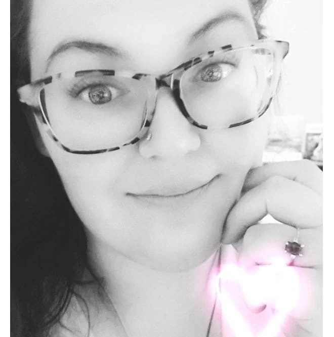 A woman with glasses smiling, holding her hand near her chin, with a bright glowing light in her hand.