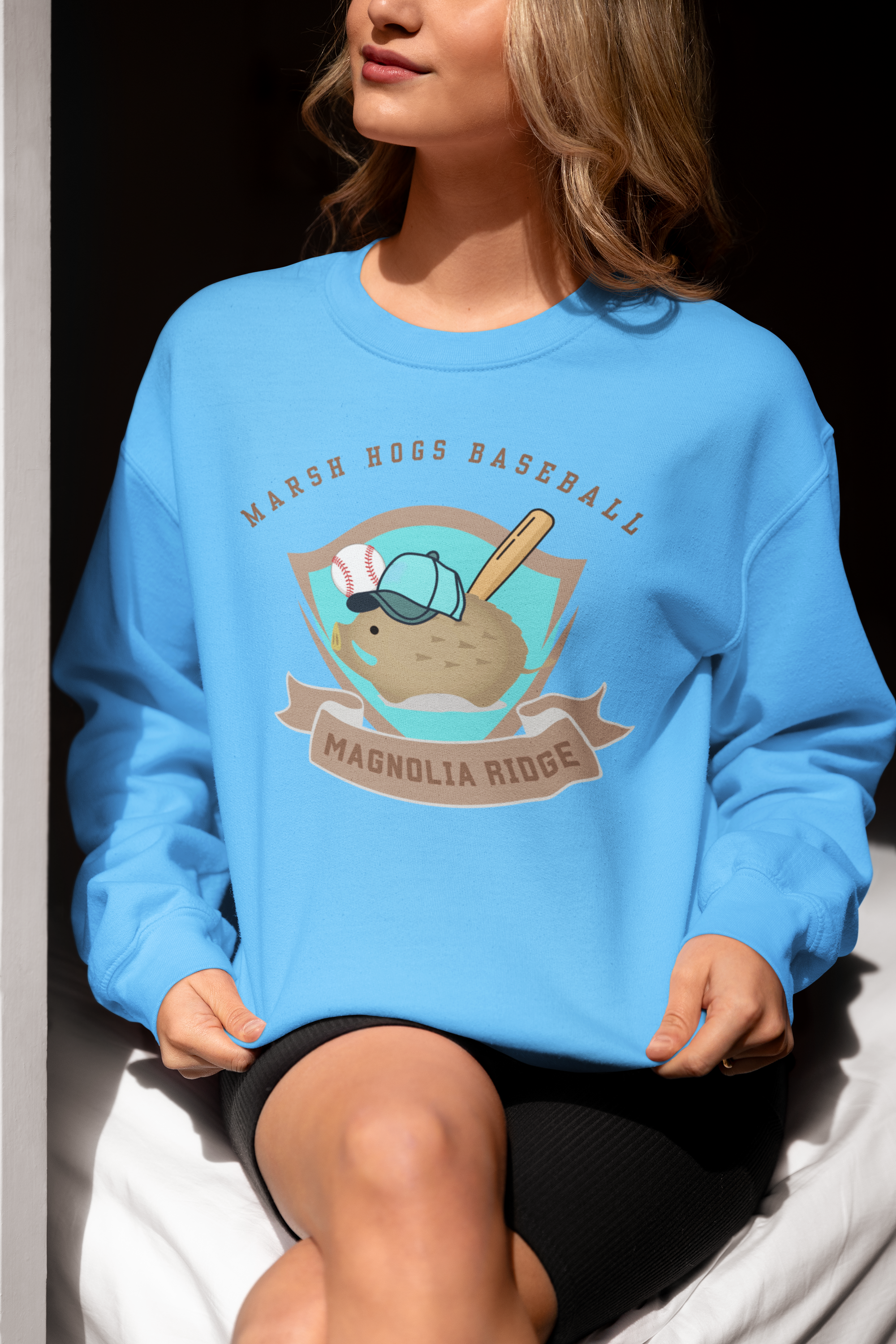 A woman wearing a blue sweatshirt with a marsh hogs baseball themed graphic on it, sitting on a bed with black shorts and one leg crossed over the other.