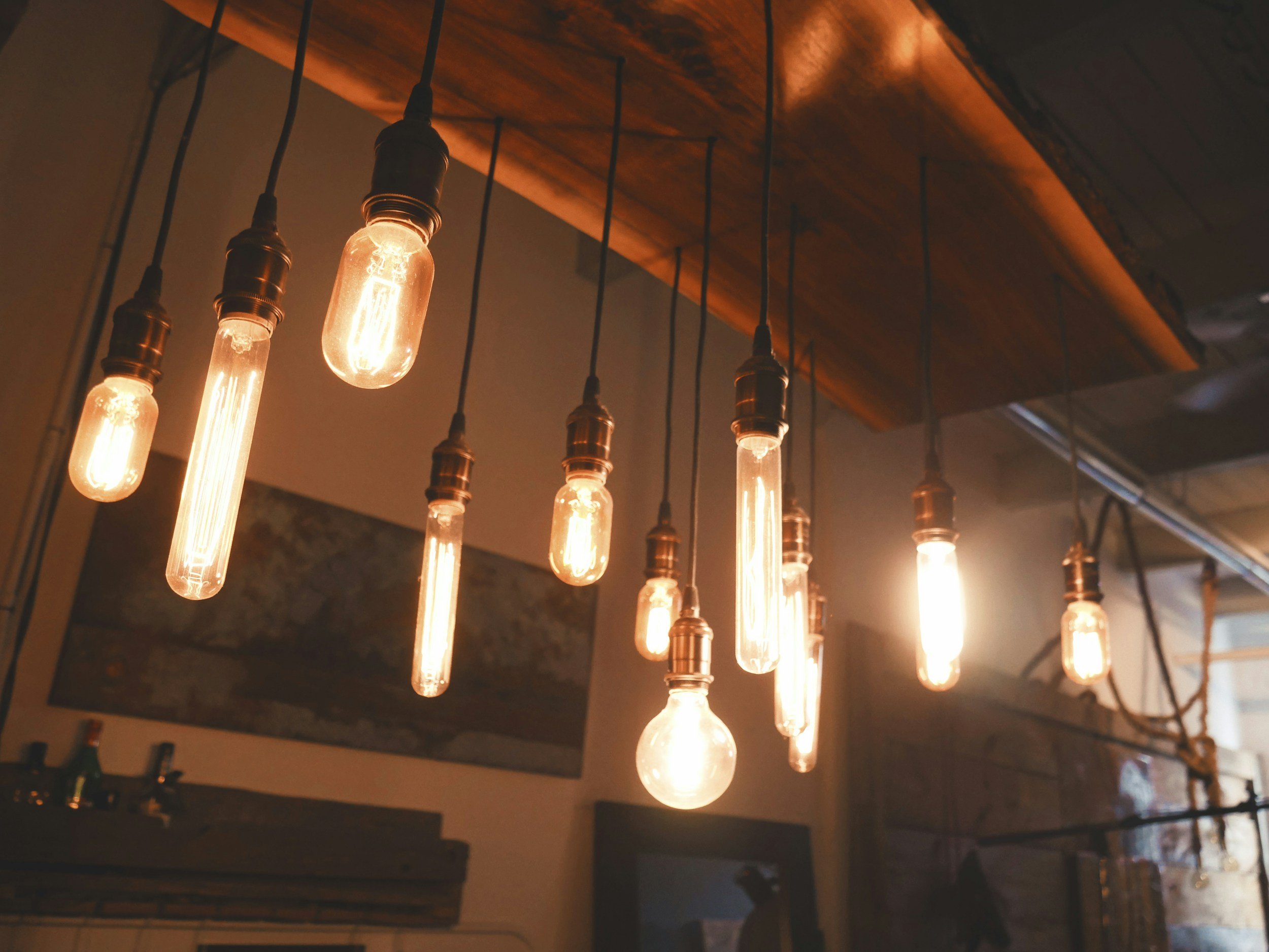 Multiple vintage-style Edison light bulbs hanging from black cords in a dimly lit room with a wooden ceiling and concrete wall.