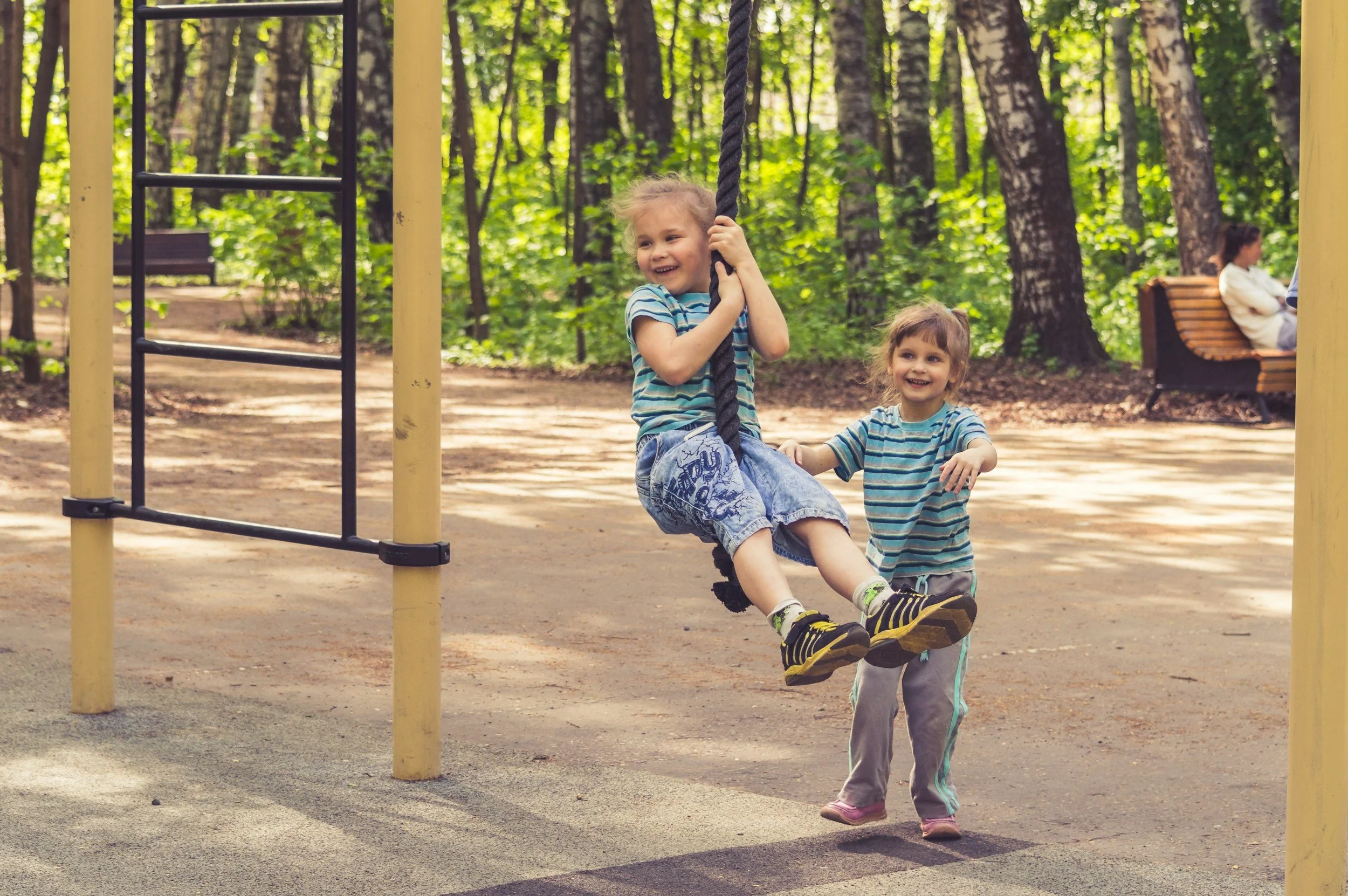 Two young girls with blonde hair wearing striped shirts, playing on a swing at a park surrounded by trees, with a woman sitting on a park bench in the background.