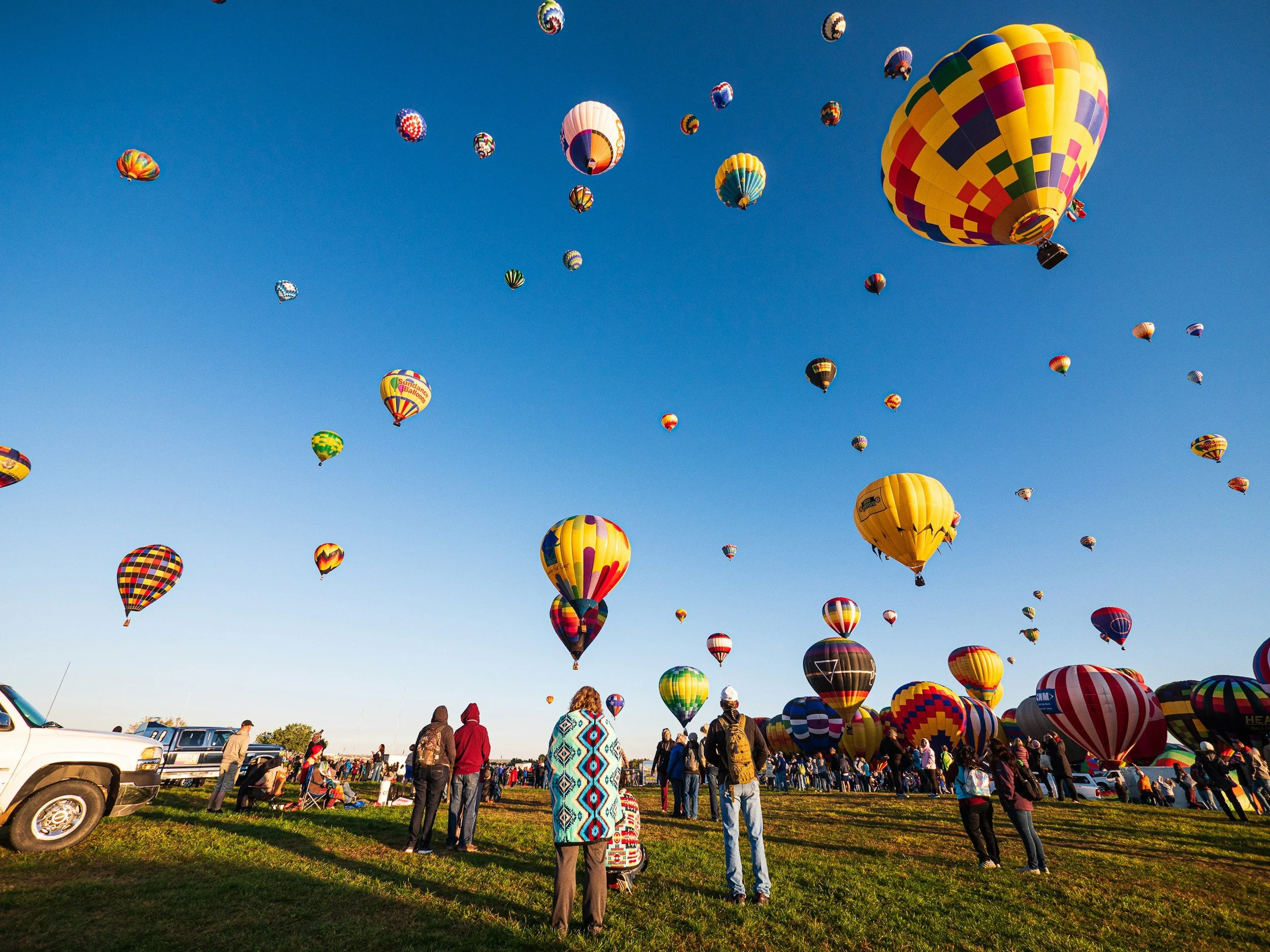 Hot air balloons in various colors and patterns flying in the clear sky while people look up and watch during daytime.