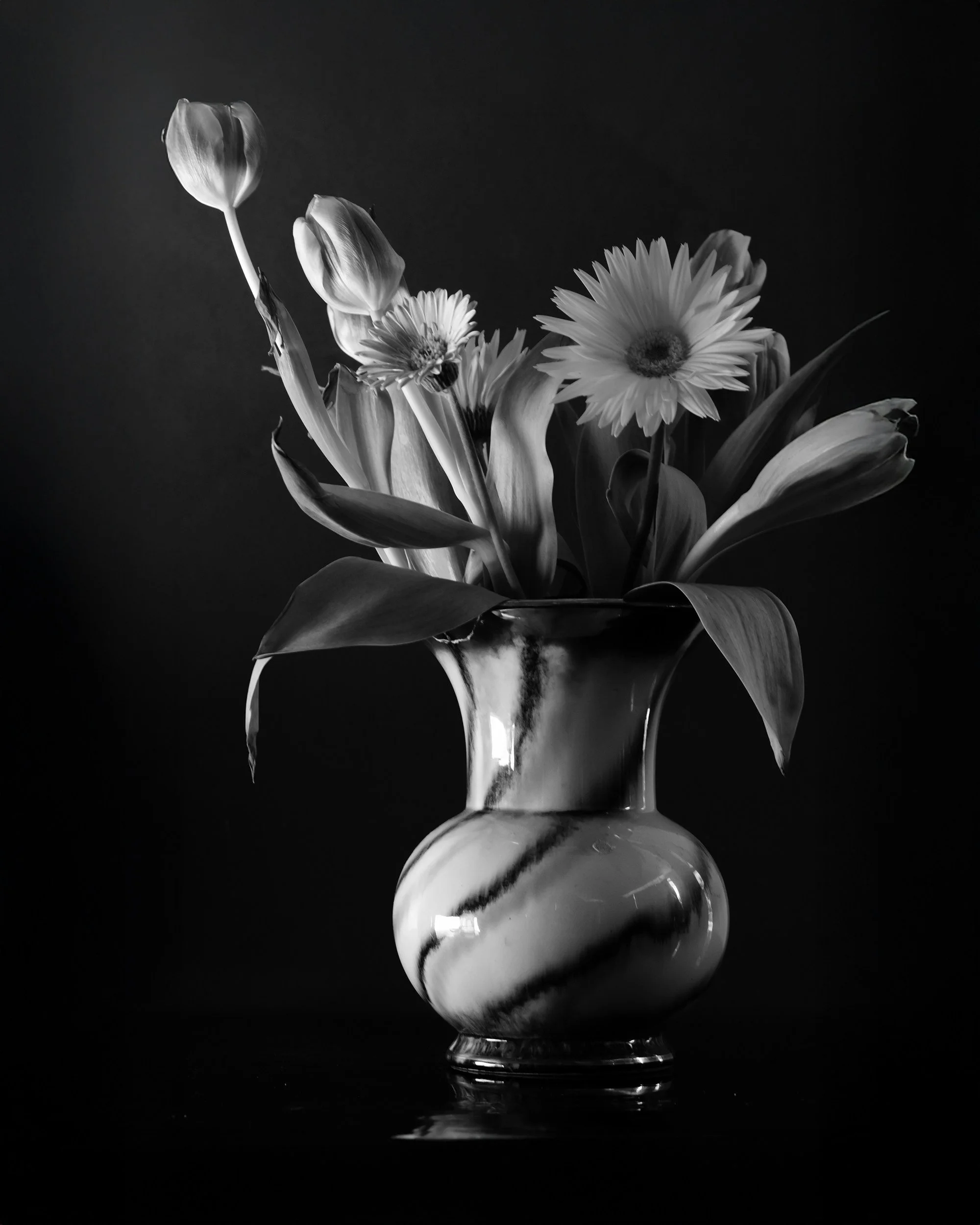 Black and white photo of a glass vase with a bouquet of flowers, including tulips and daisies, set against a dark background.