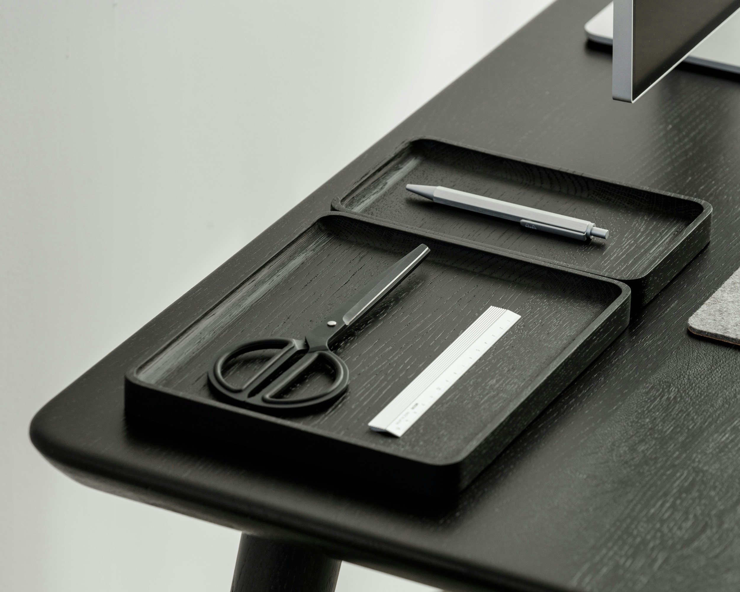 Black desk with a white stylus pen in a tray, scissors, and a silver monitor stand.