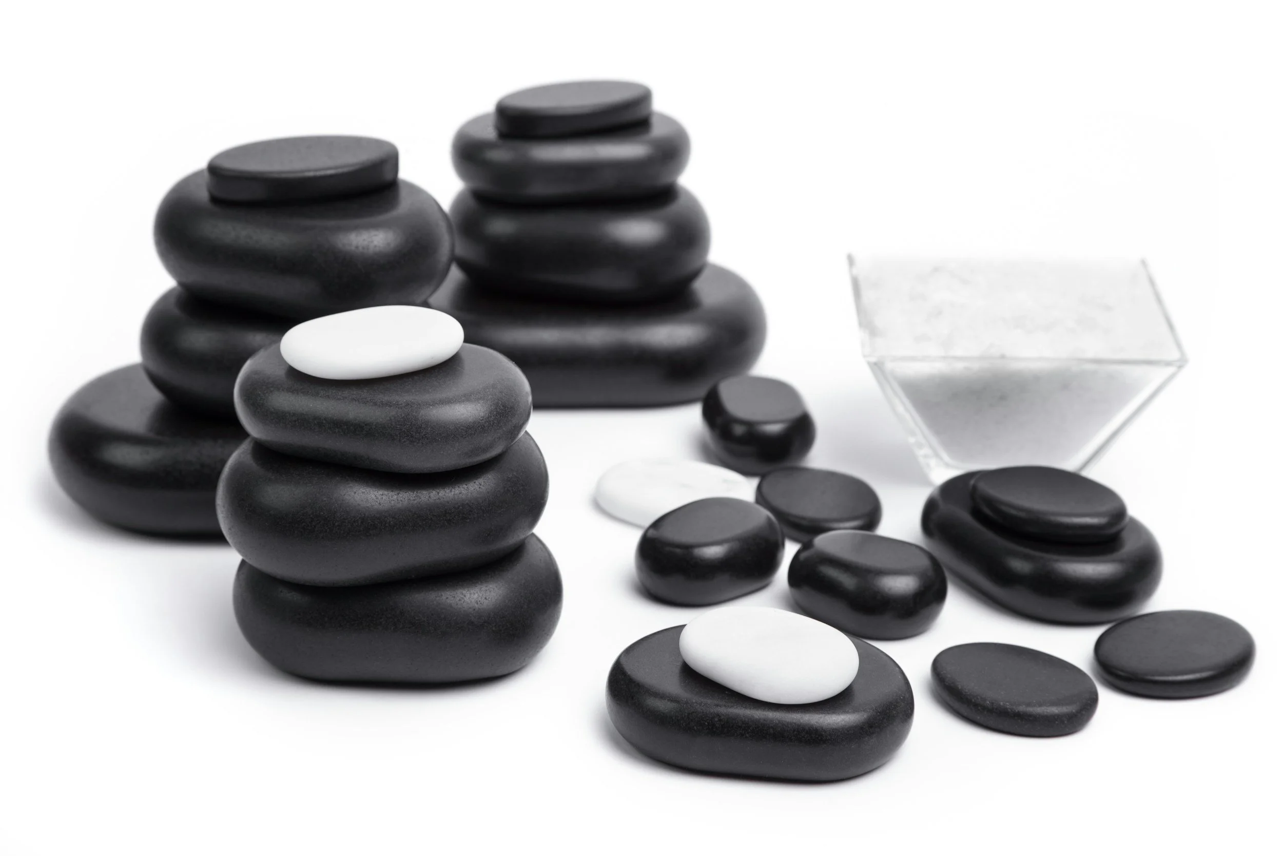 Stacked black stones, some with smaller stones on top, with white stones among them, and a clear glass container nearby on a white background.