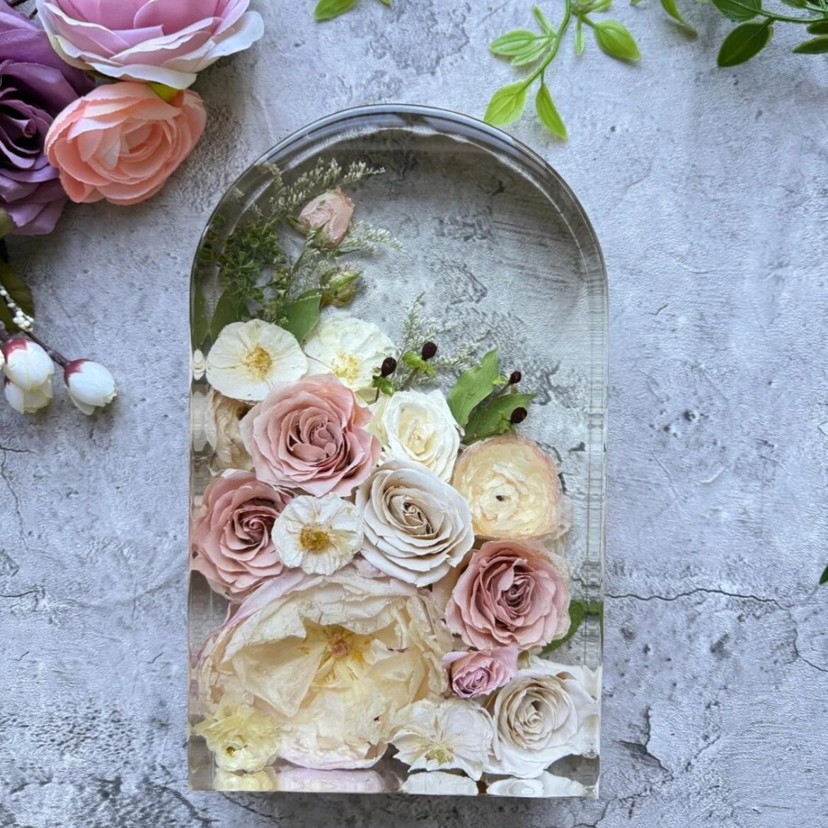 A glass container filled with floating pastel-colored roses and flowers, placed on a gray textured surface with additional flowers and green leaves nearby.
