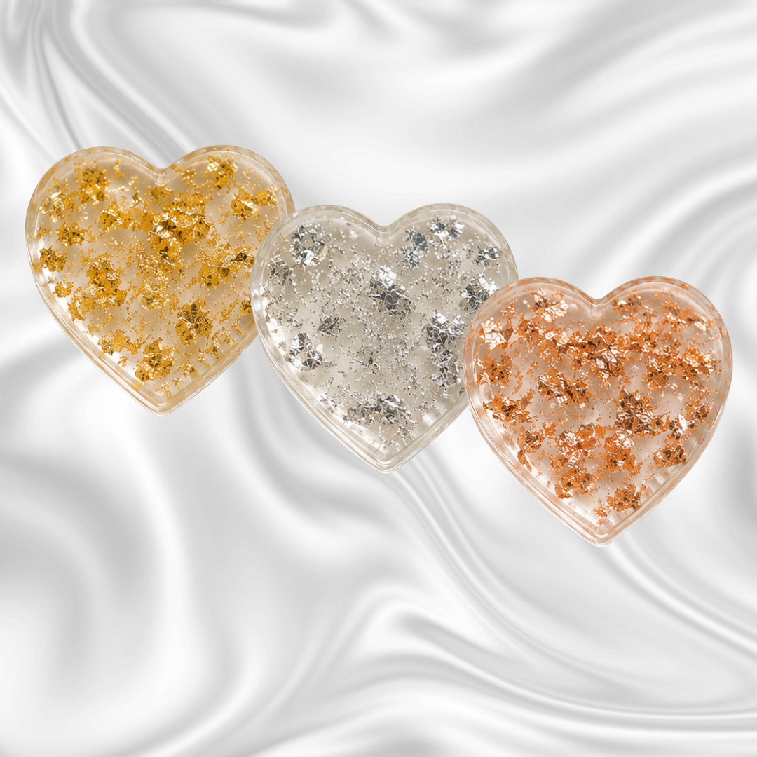 Three heart-shaped containers filled with glitter, in gold, silver, and rose gold colors, arranged on a dark, swirled background.