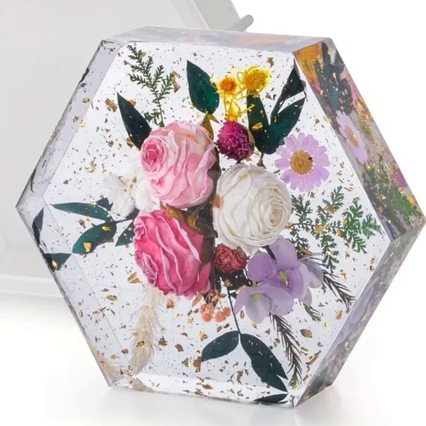 A clear, hexagonal resin paperweight filled with pink, white, and purple pressed flowers and green leaves, with gold flakes embedded inside.