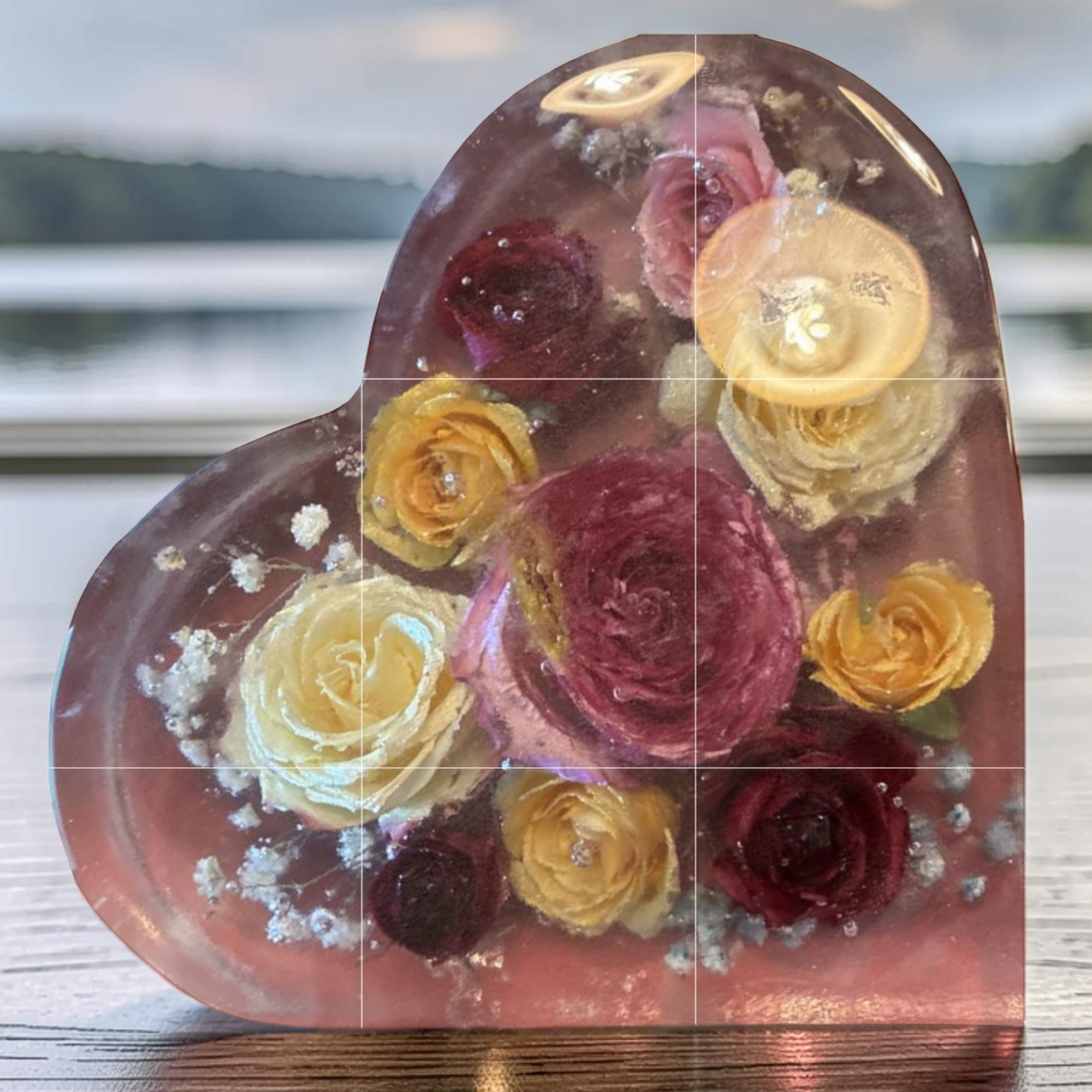 A heart-shaped glass paperweight with embedded colorful flowers resting on a wooden surface with a blurred landscape background.