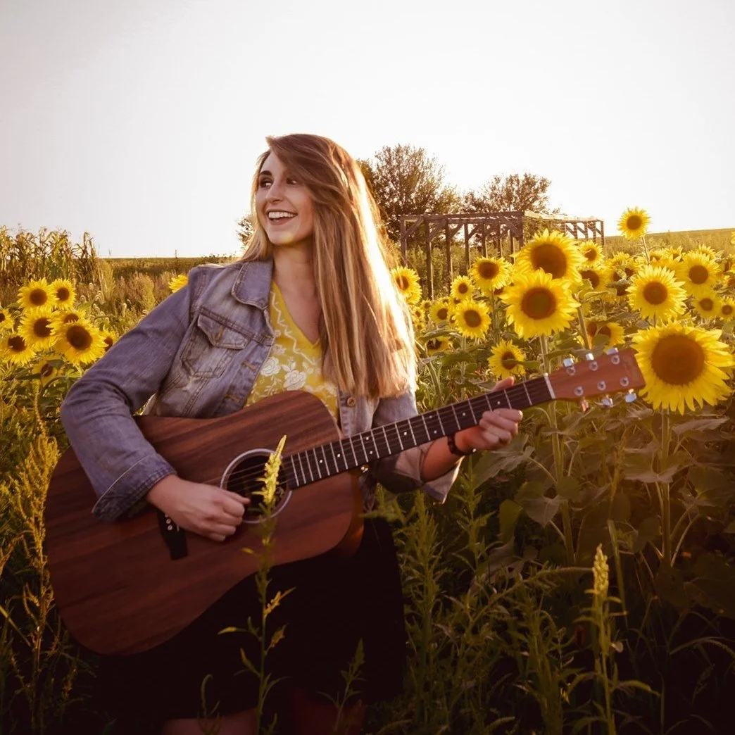 gvb marketing agency employee, Emily Hoving, playing an acoustic guitar in a sunflower field during sunset
