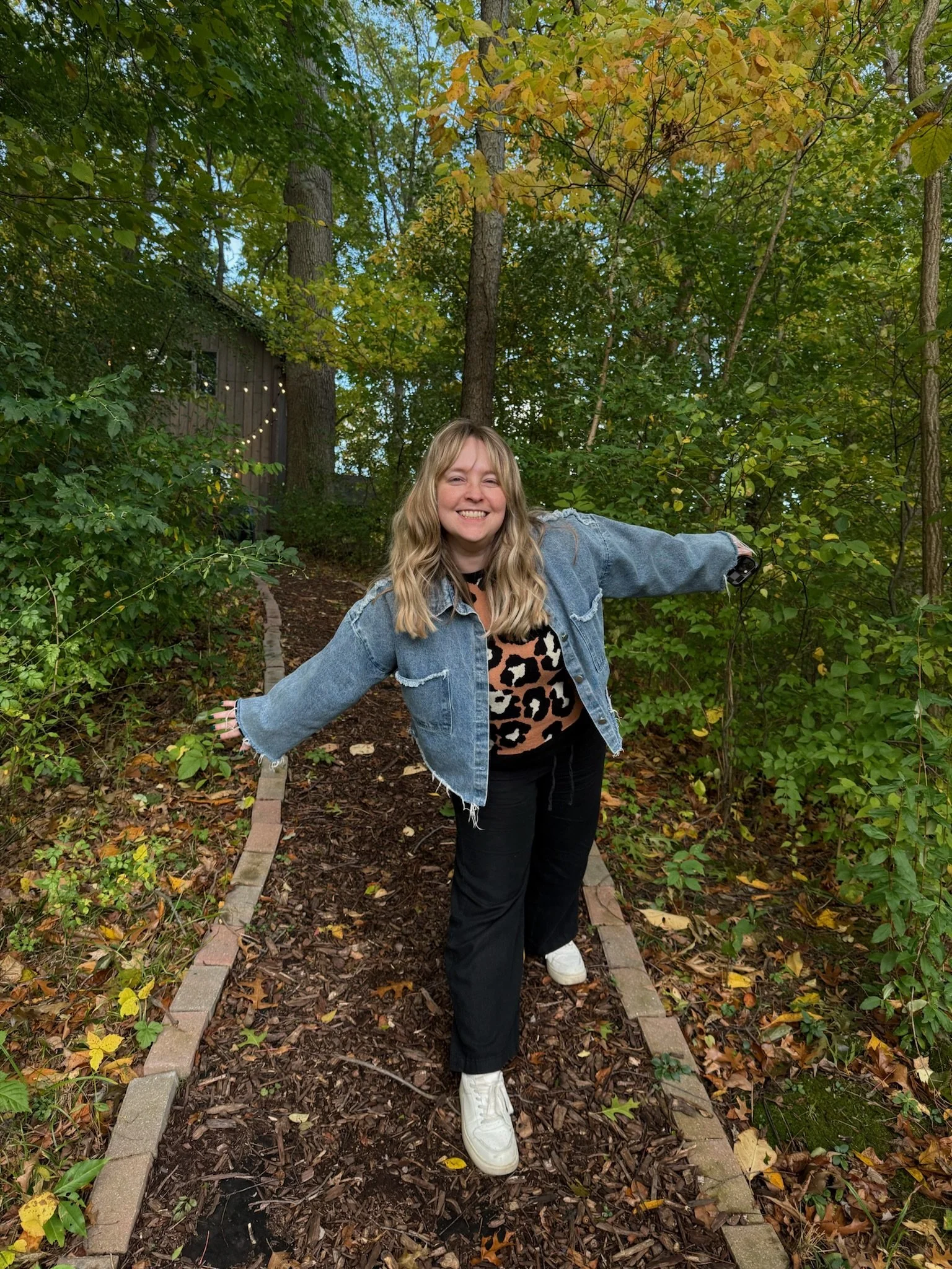 gvb marketing agency employee, Kelsey Johnson, smiling and posing with arms outstretched on a wooded path with trees and autumn leaves.