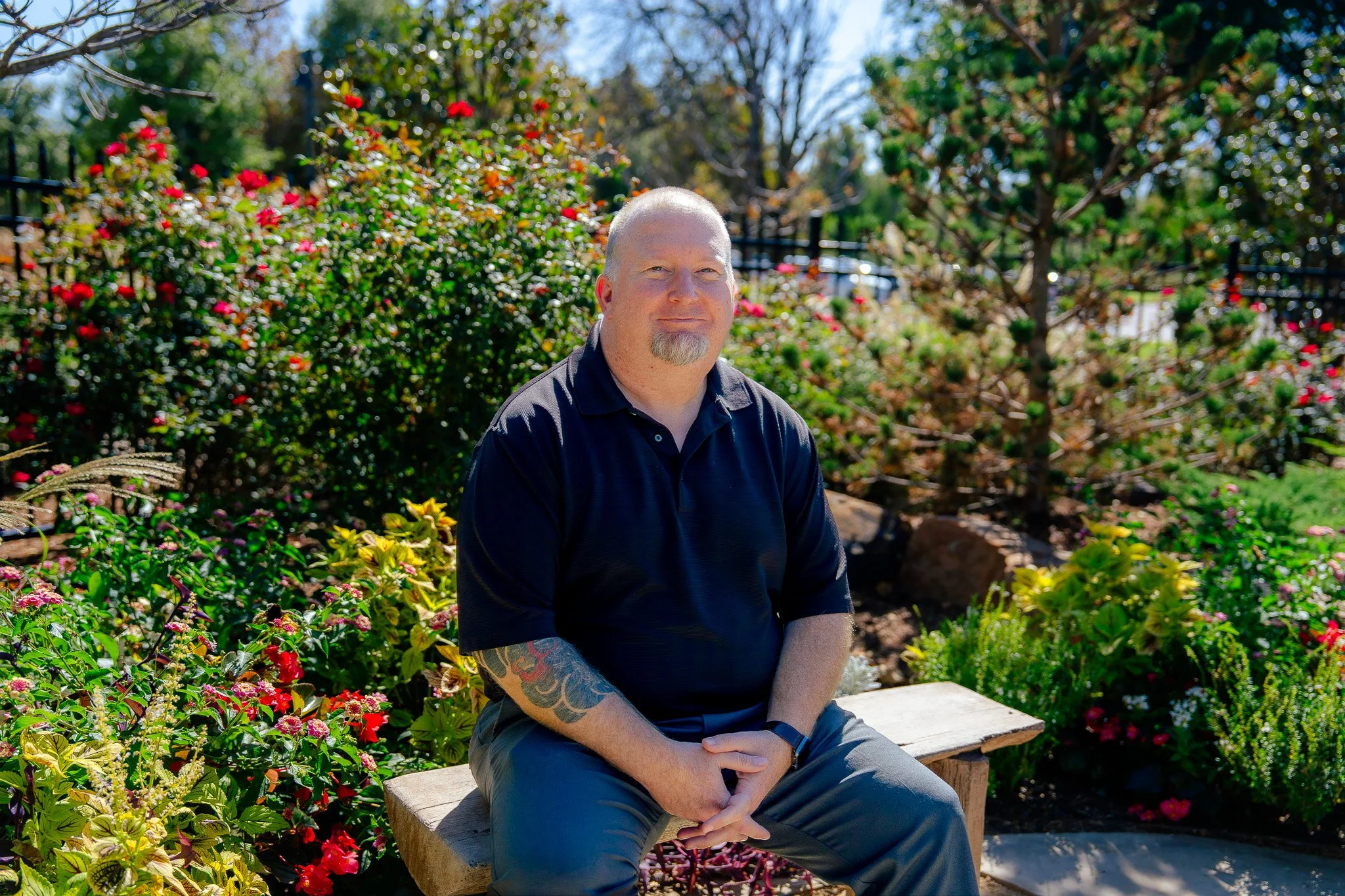 A man with a shaved head and goatee, wearing a black polo shirt and gray pants, sitting outdoors on a stone bench in a garden with colorful flowers and trees in the background.