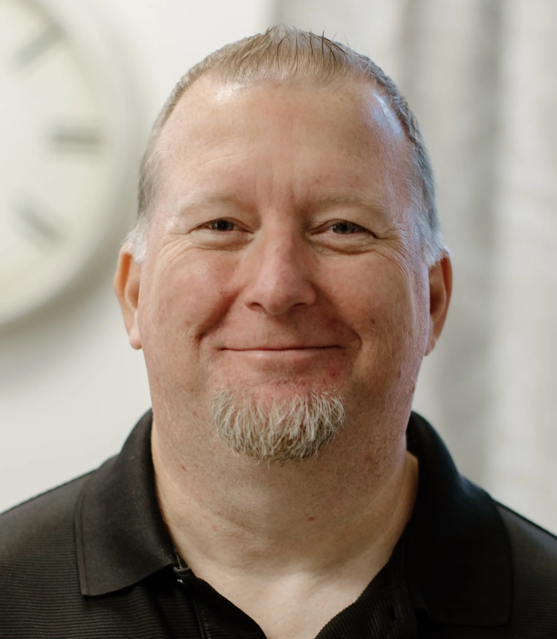 Close-up of a smiling middle-aged man with short blond and gray hair, beard, wearing a black shirt, against a light-colored background.