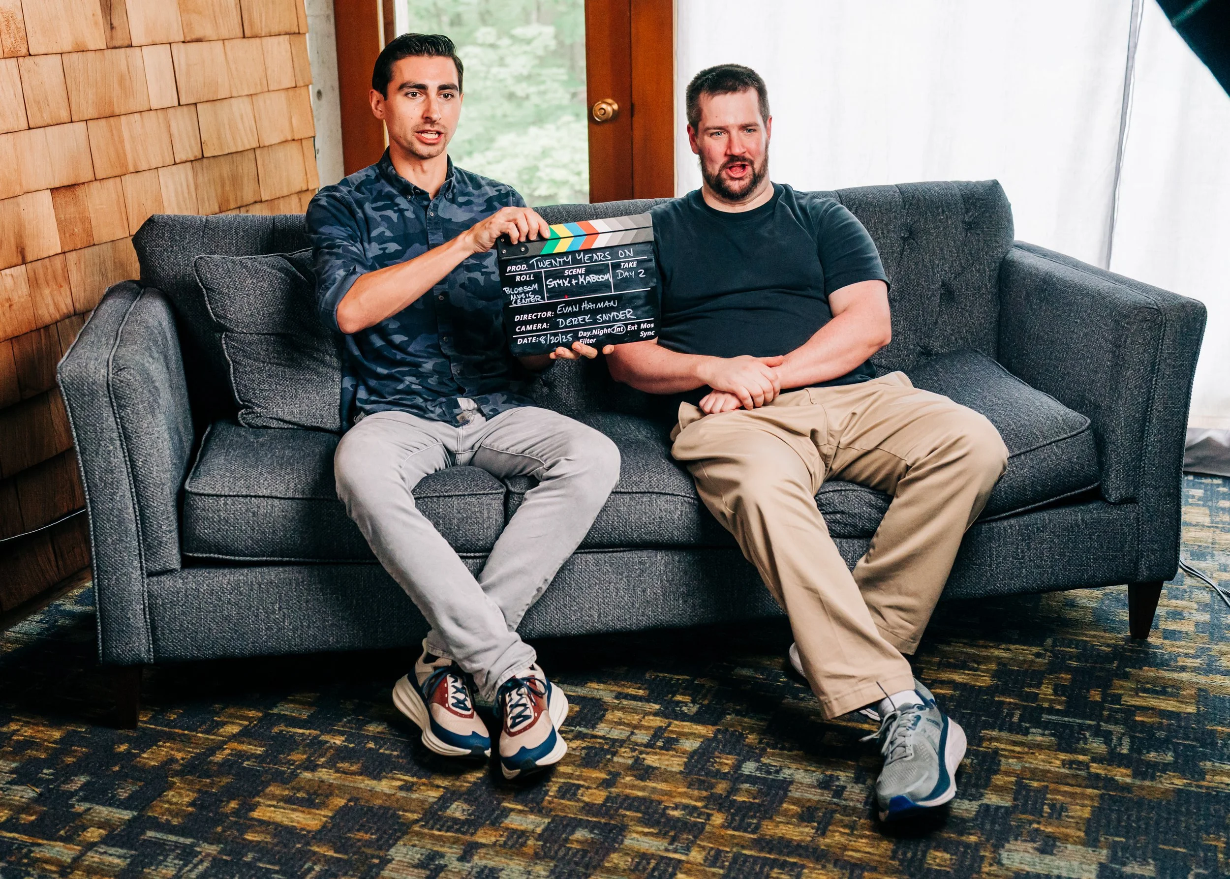 Two men sitting on a gray couch in a room with wood-paneled wall and large window. One man holds a clapperboard with production details, both men are engaged in a conversation.