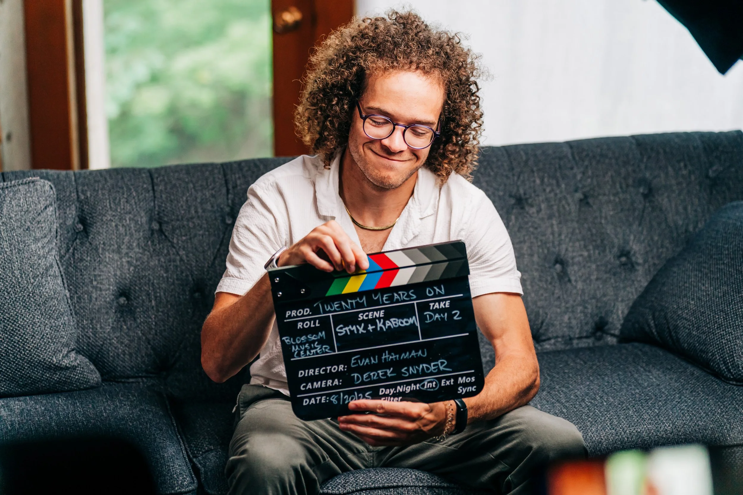 A man with curly hair and glasses sitting on a gray couch holding a film slate with production details, smiling.