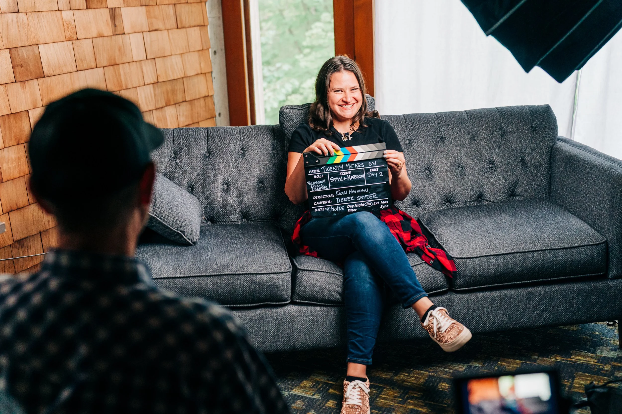 A woman smiling while sitting on a gray couch holding a clapperboard during a video or photo shoot. Two people are partially visible in the foreground, one with dark hair and the other with a hat. The setting appears to be indoors with natural light and wooden wall paneling.