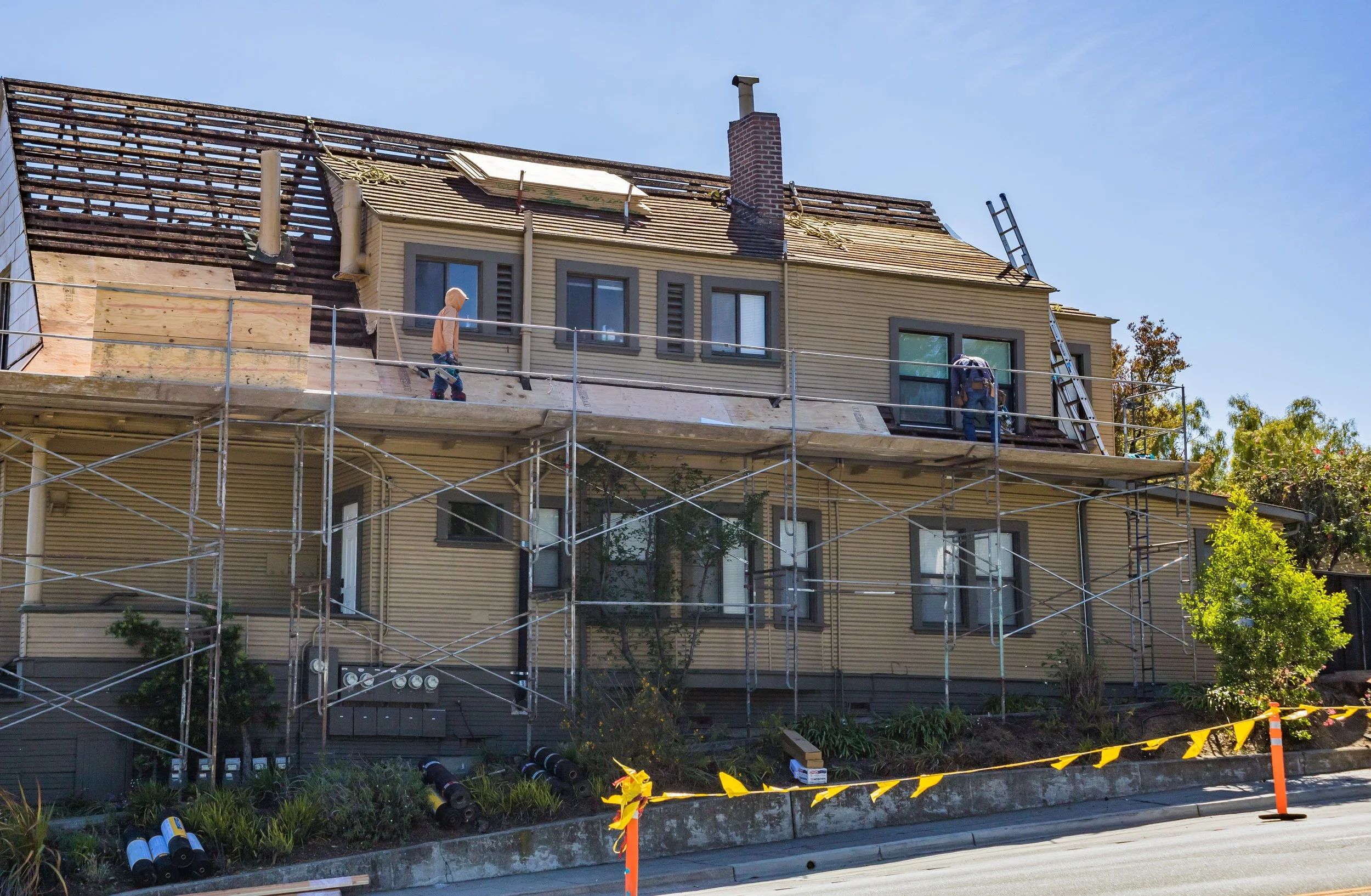 Two workers on scaffolding repairing the roof of a tan, multi-story house with large windows, with trees and a blue sky in the background.