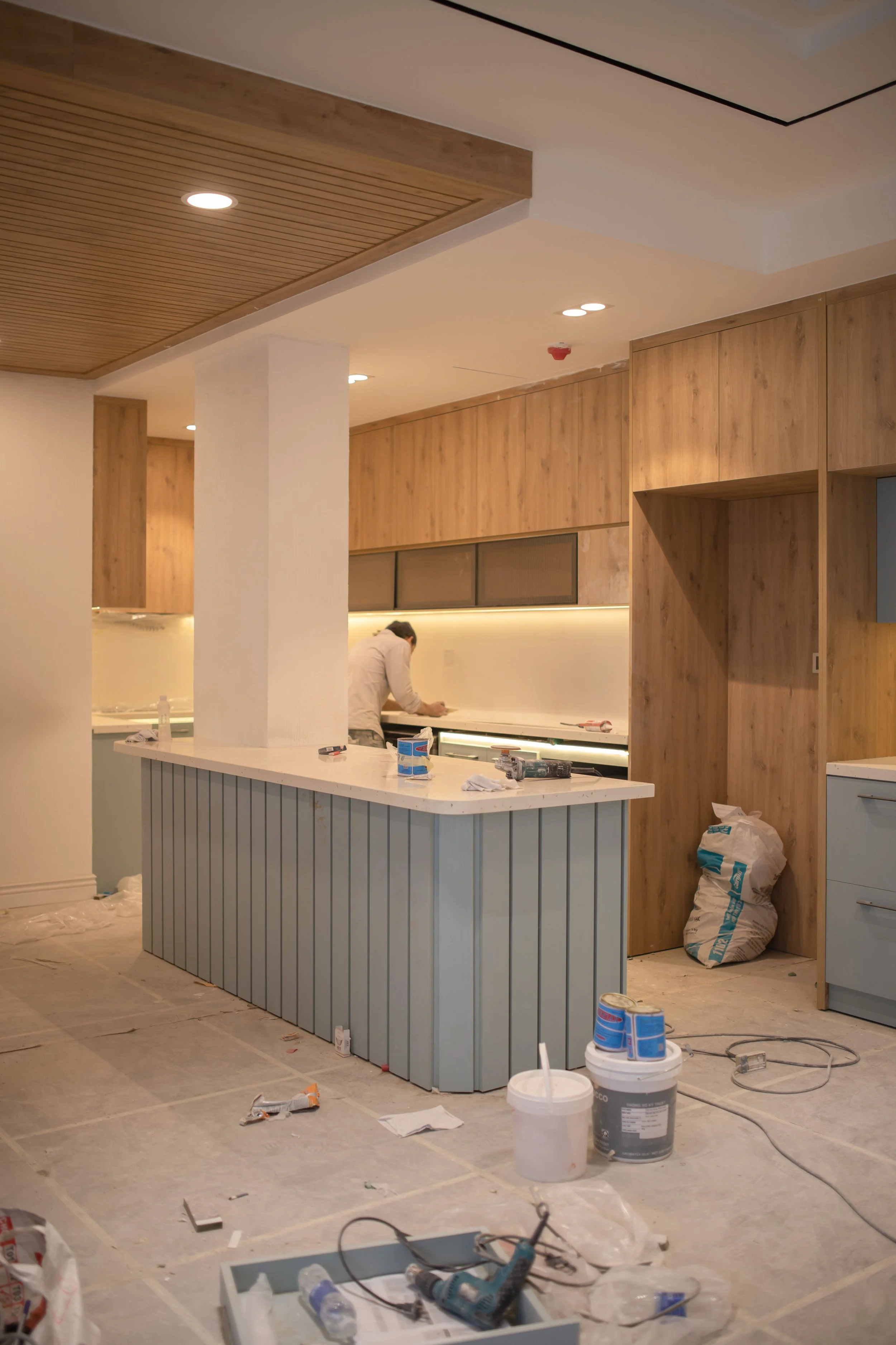 Interior of an unfinished kitchen under construction with a light blue distressed wood bar counter, wooden cabinets, and construction tools and supplies scattered on the floor.