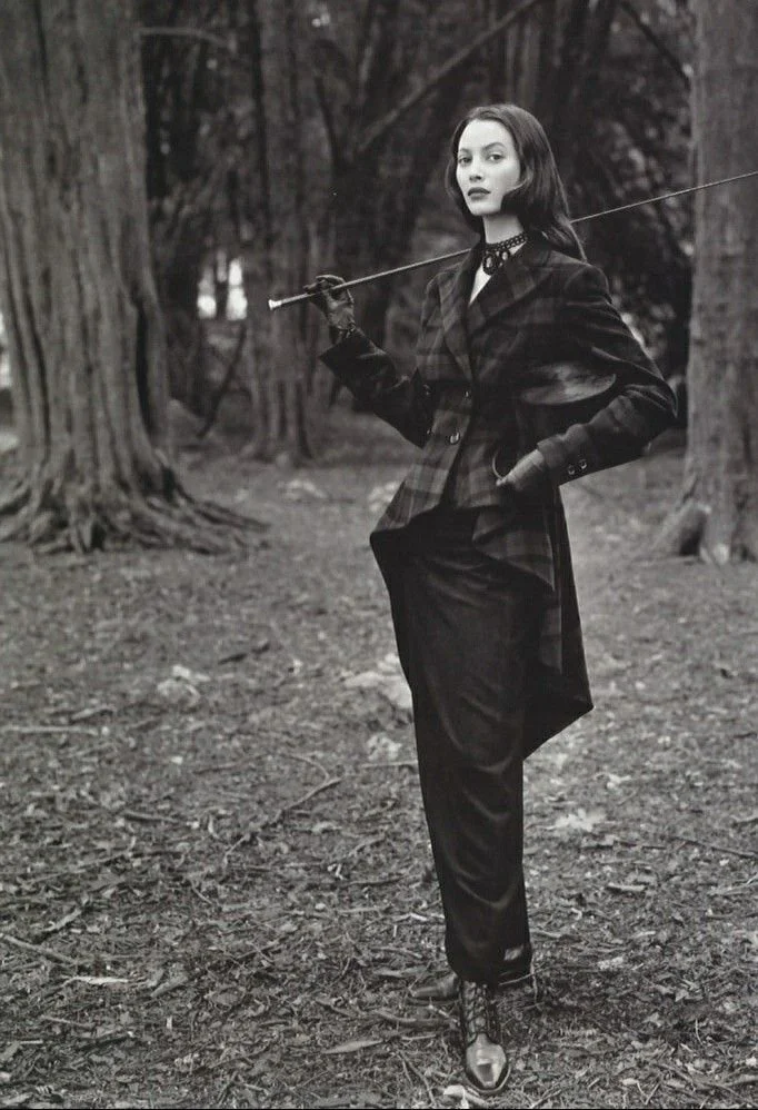 Model Christy Turlington dressed in dark, stylish British clothing stands in a wooded area holding a rod across her shoulders. She has shoulder-length hair and is wearing gloves, a choker, and lace-up boots.