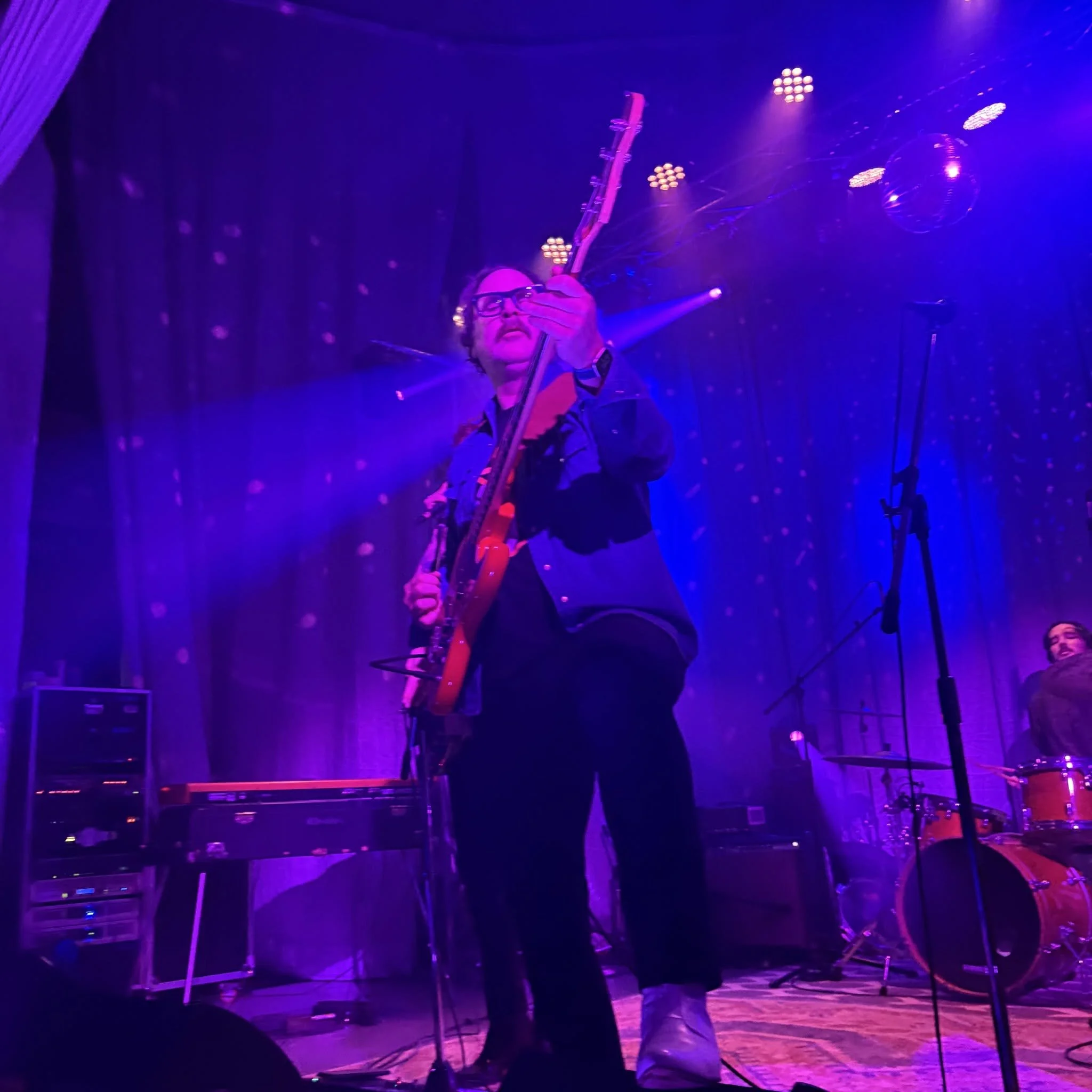 A musician with glasses and a dark jacket playing an electric guitar on stage illuminated by purple and pink stage lights, with reflections and spotlights in the background.