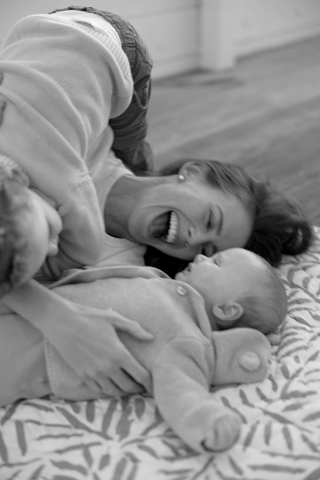 A woman and two children lying on a bed, smiling and playing together.