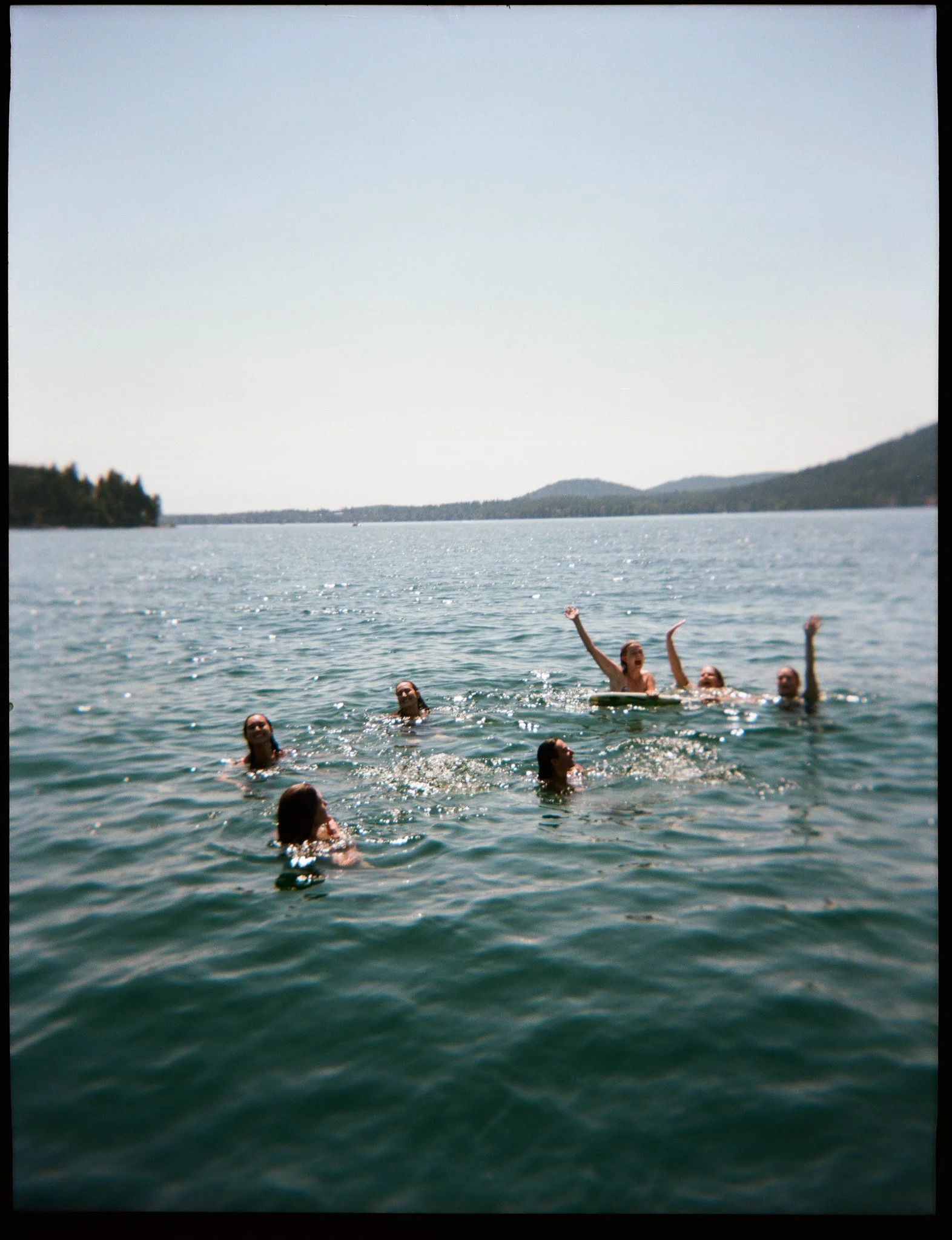Group of people swimming and playing in a body of water with hills in the background and a clear sky.