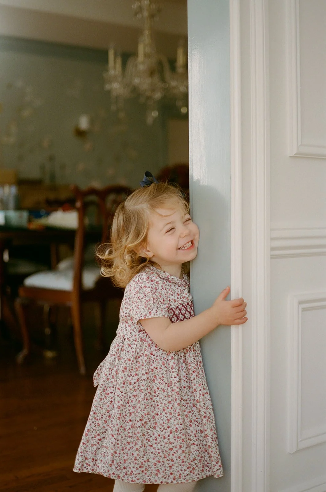 A young girl with curly blonde hair, wearing a floral dress, happily peeking from behind a white door.