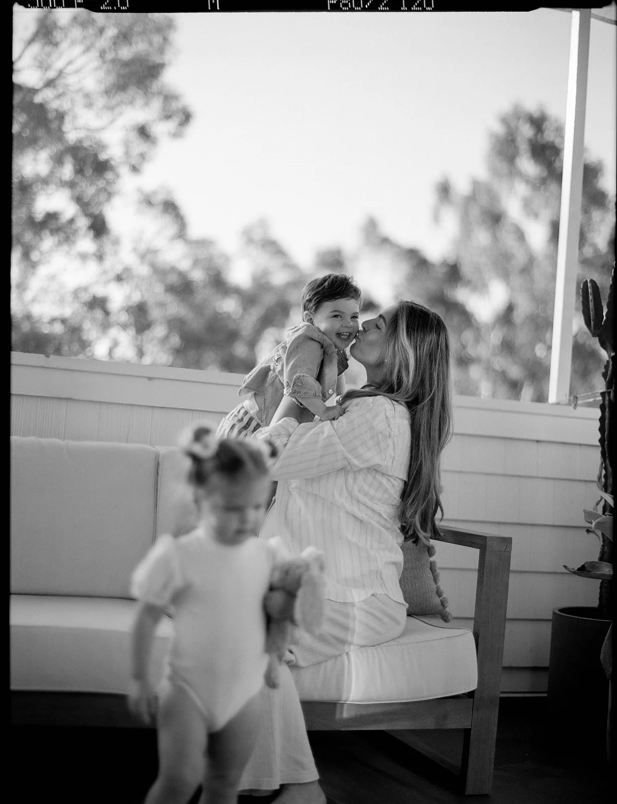 A woman sitting on a couch holding a young child in her arms, both smiling and leaning in for a kiss. A blurred child stands in the foreground, and a window with trees outside is visible in the background.
