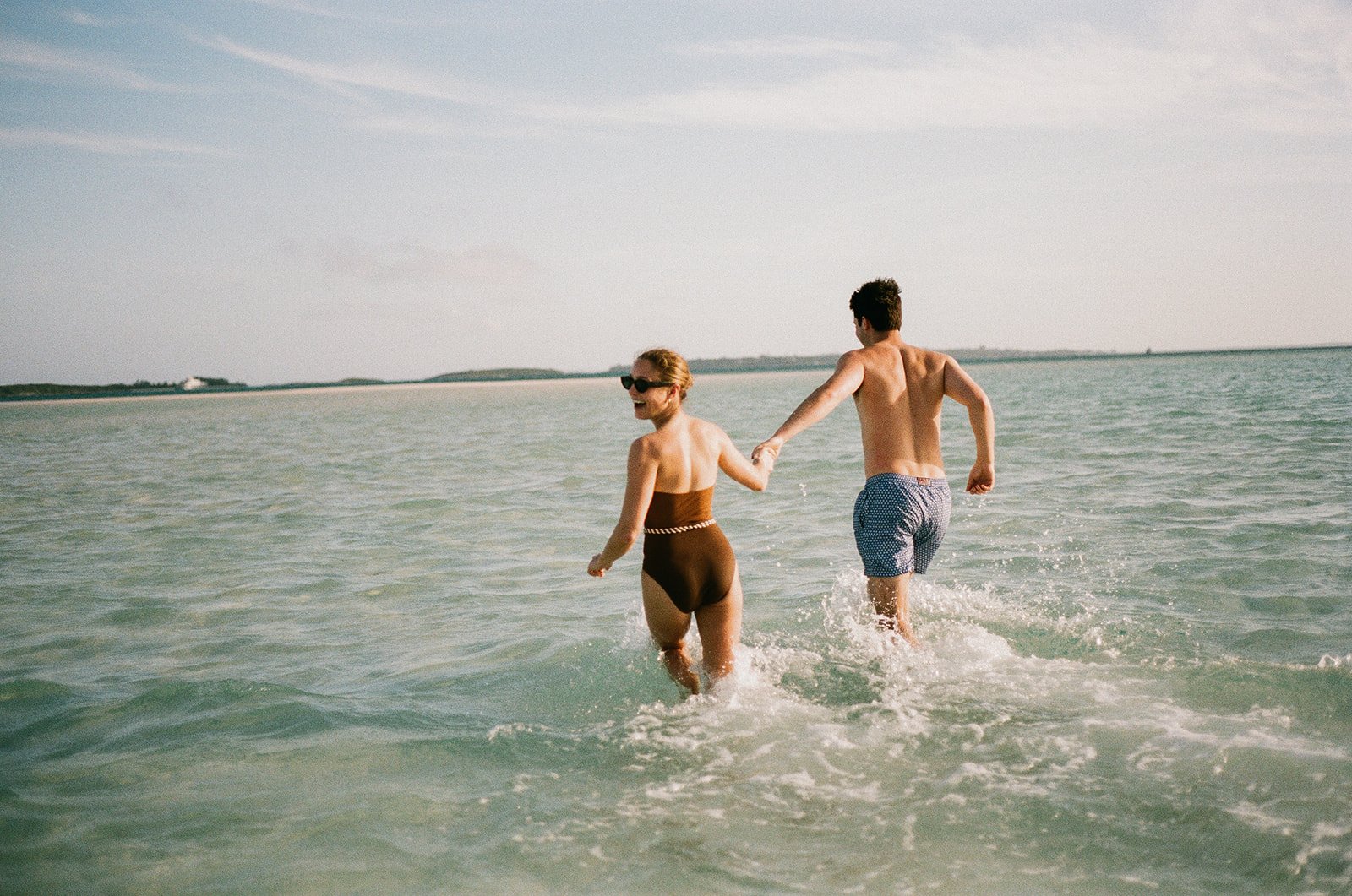 A man and a woman holding hands and walking in the ocean, wearing swimsuits, with the woman laughing and wearing sunglasses. There are distant landmasses on the horizon under a clear sky.