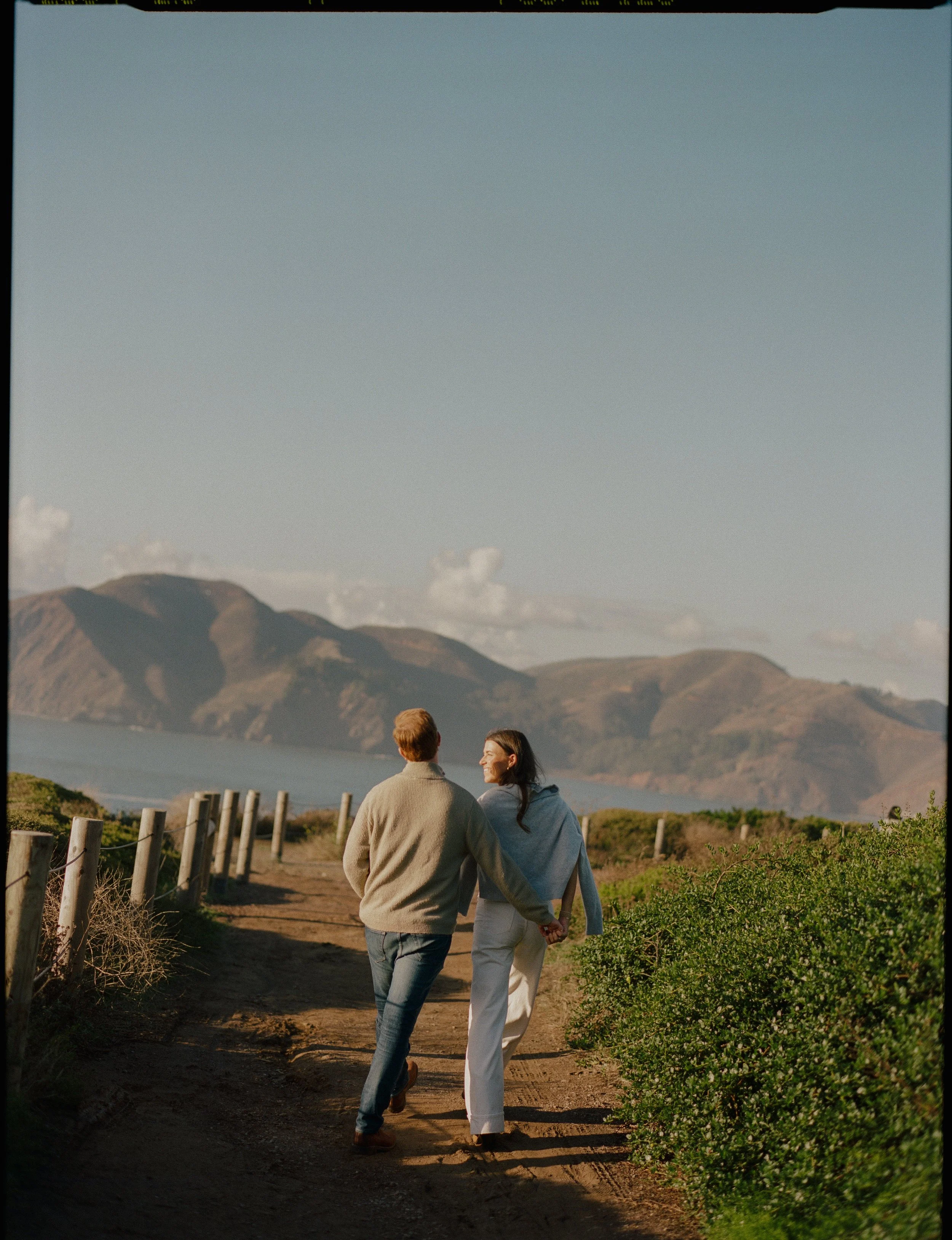 A couple walking hand in hand along a dirt path near water, with hills and mountains in the background, during what appears to be late afternoon or early evening.