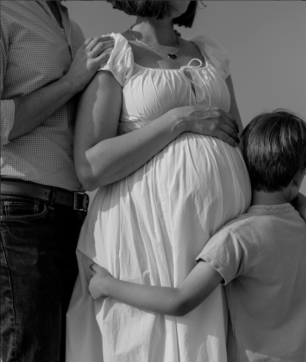 A pregnant woman in a white dress is surrounded by her partner and son, holding her belly in an intimate family moment.