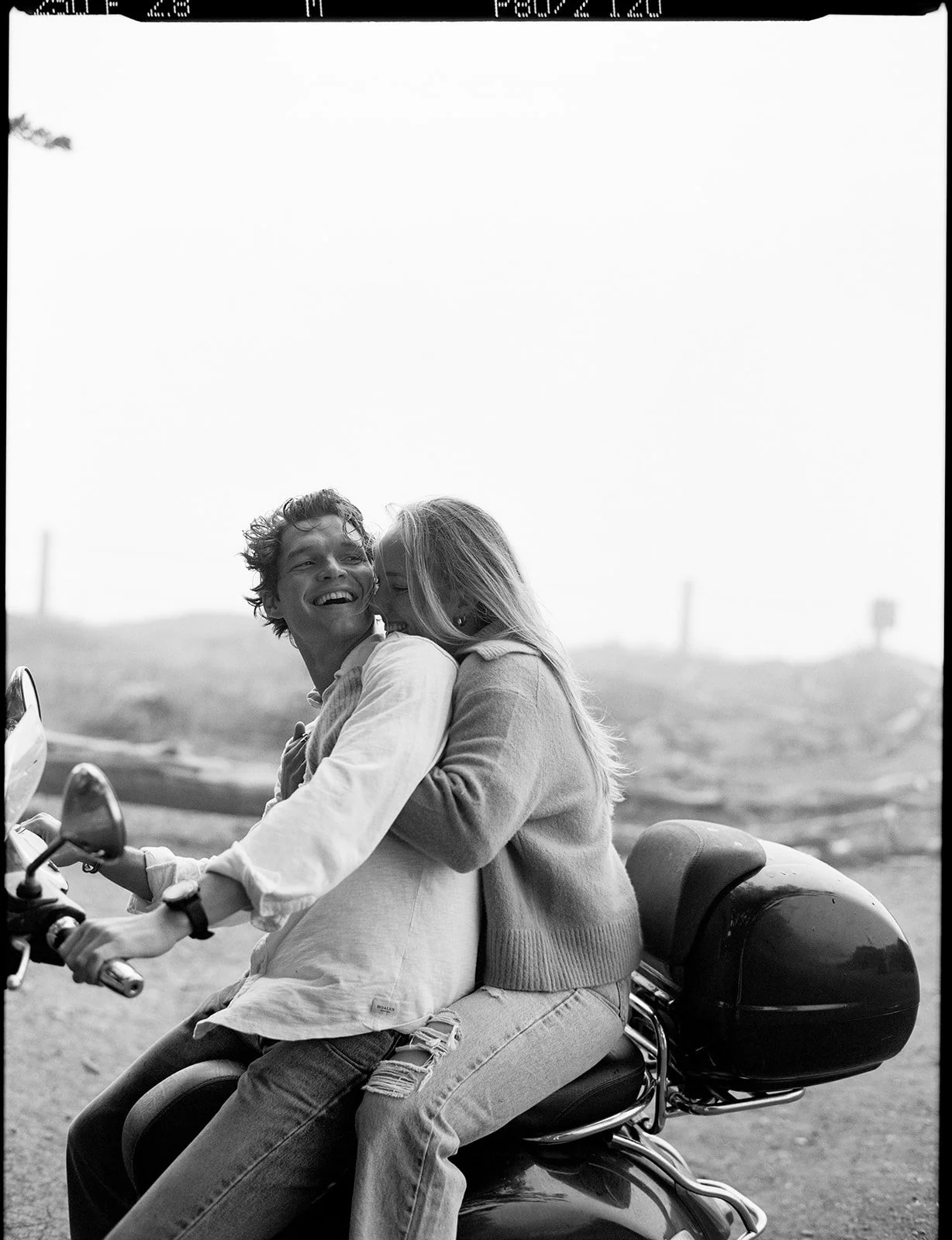 A black-and-white photo of a young man and woman riding a motorcycle together outdoors. The woman is whispering into the man's ear, and they are both smiling and appear joyful.