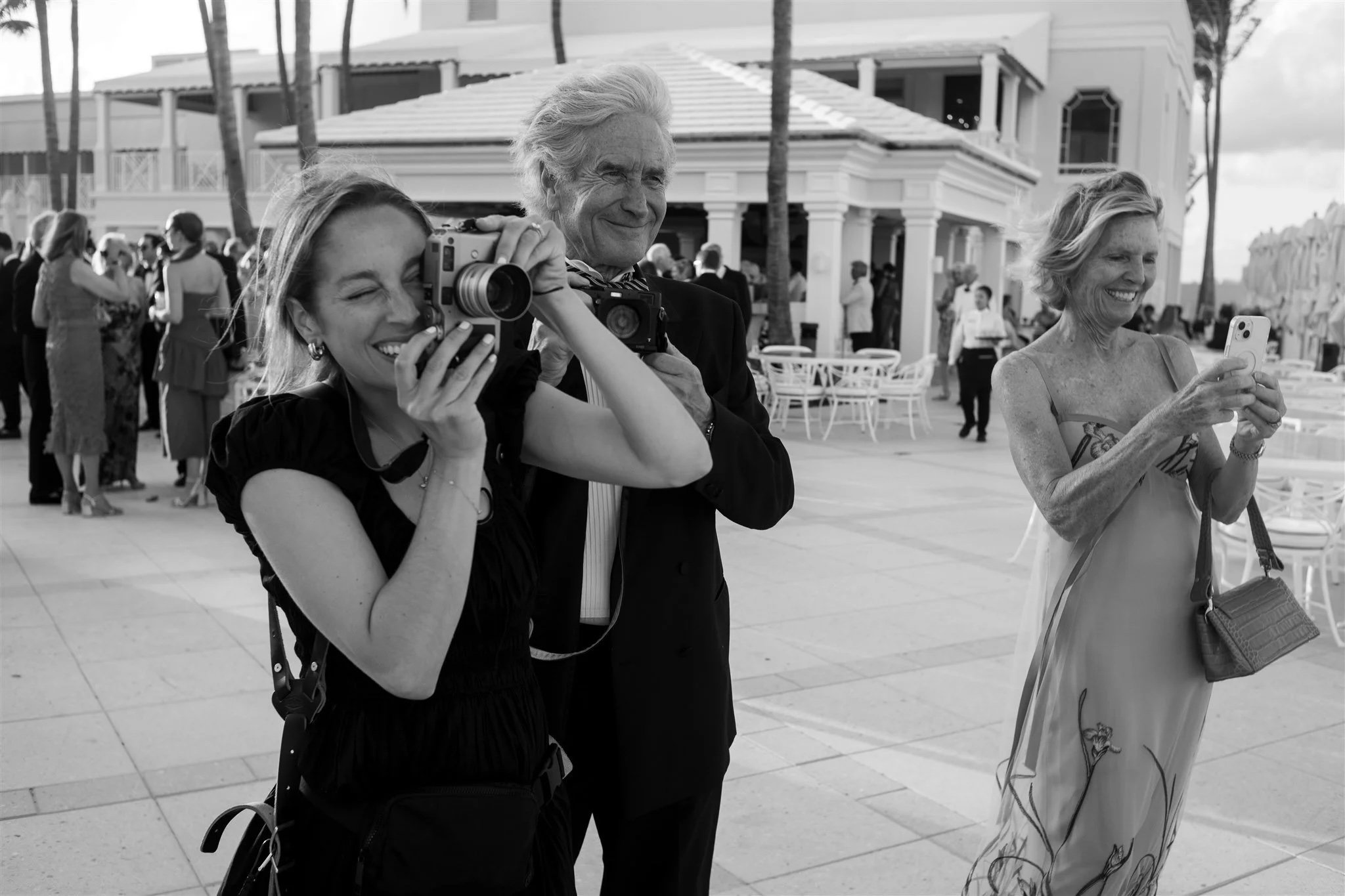 Three people at an outdoor event: a woman with long hair taking a photo or video with a camera, an older man with gray hair smiling, and a woman with short hair looking at her phone, all in front of a large building and surrounded by other attendees.