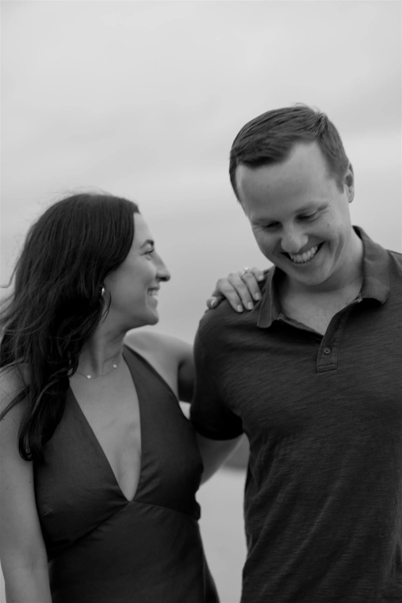 A smiling woman and man with their heads tilted towards each other, standing closely outdoors on a cloudy day.