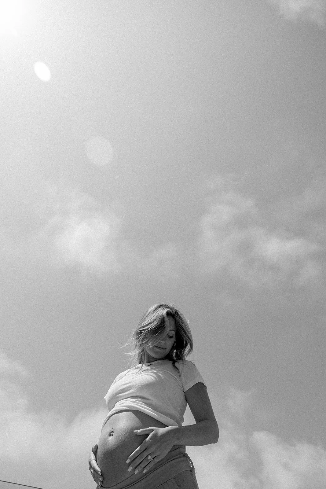 Black and white photo of a pregnant woman standing outside, smiling while holding her belly. The sky with some clouds is visible in the background.