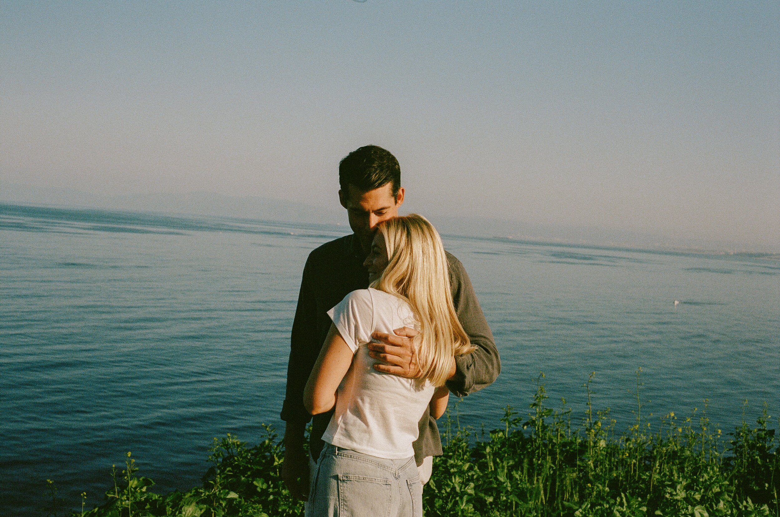 A man and a woman hugging by a lake at sunset, with the water and sky visible in the background.