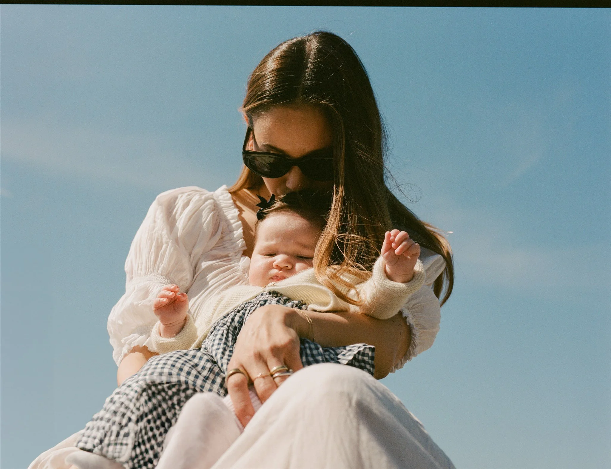 A woman with brown hair wearing large black sunglasses and a white blouse holds a smiling baby with dark hair, a small black bow in her hair, and a beige sweater, against a clear blue sky.