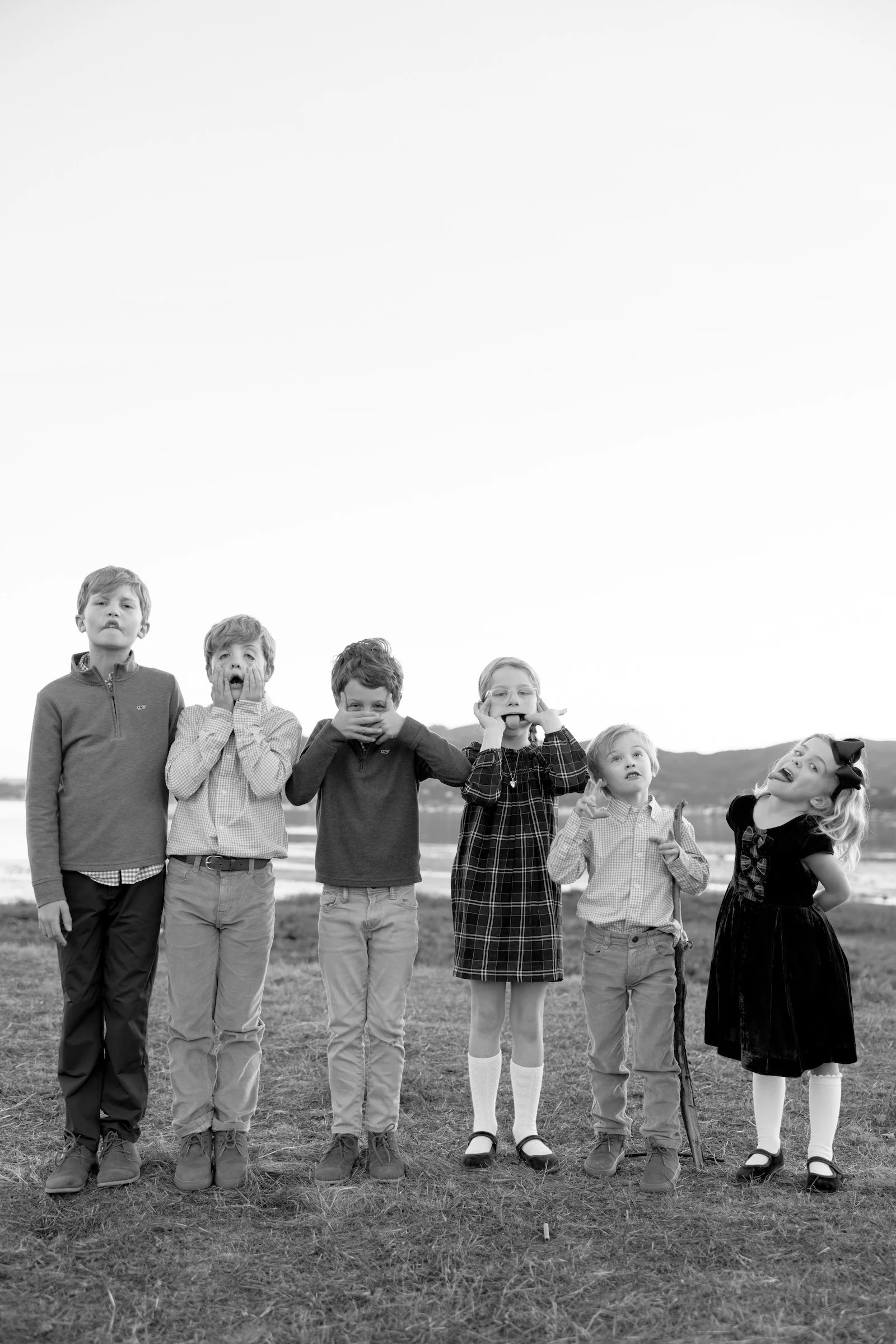 Black and white photo of seven children standing outdoors on grass with mountains and sky in the background. The children display various facial expressions, some surprised, some playful, some serious, and one with her tongue sticking out.