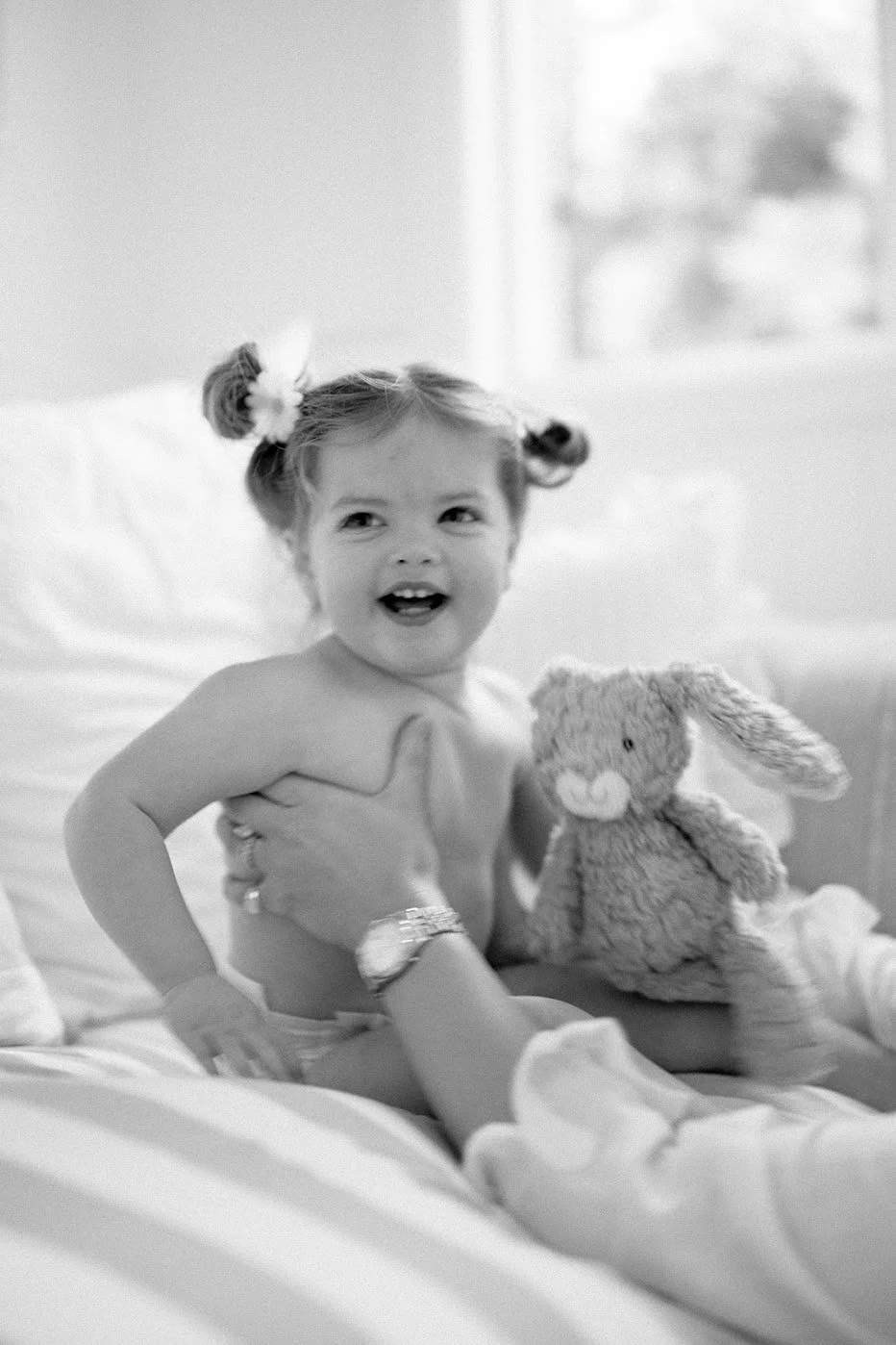A young girl with braided hair sitting on a bed, holding a stuffed bunny, smiling and looking to the side, with a window in the background.